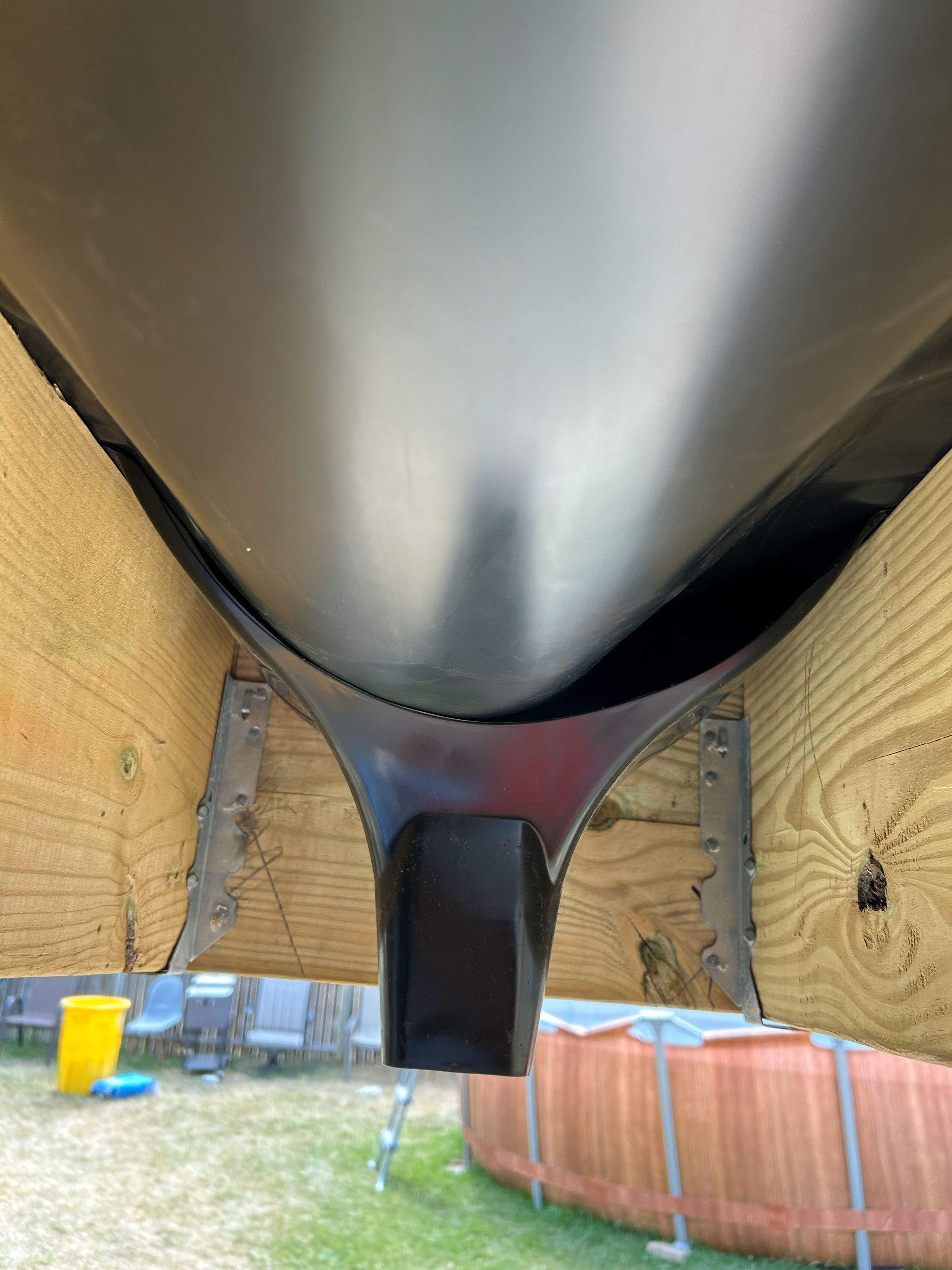 Black gutter with downspout, supported by wooden beams, overlooking a backyard with an above-ground pool.