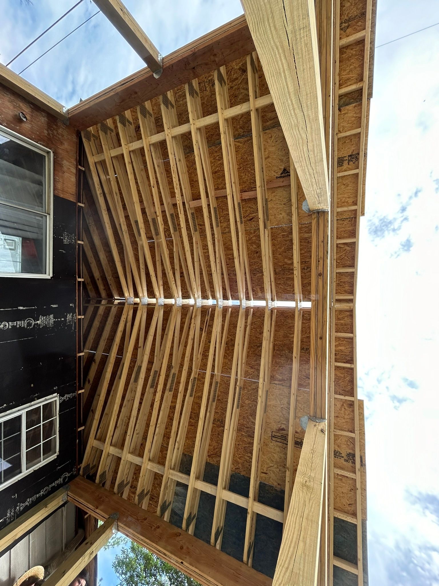 Framing of a roof in construction, with wooden beams and sheathing. Black material on walls, sky visible.