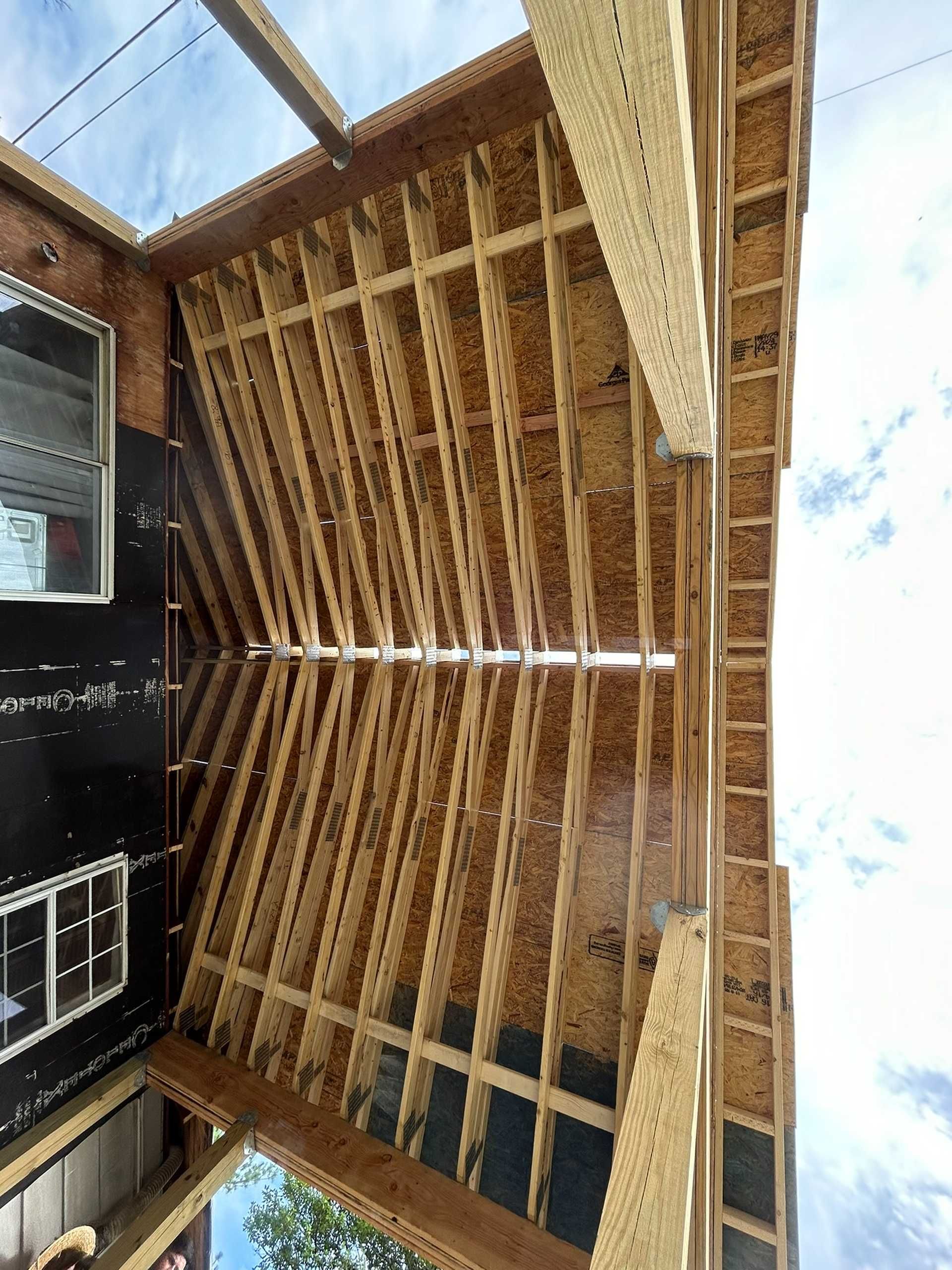 Construction: Wooden roof framing with black sheathing and visible rafters against a blue sky.