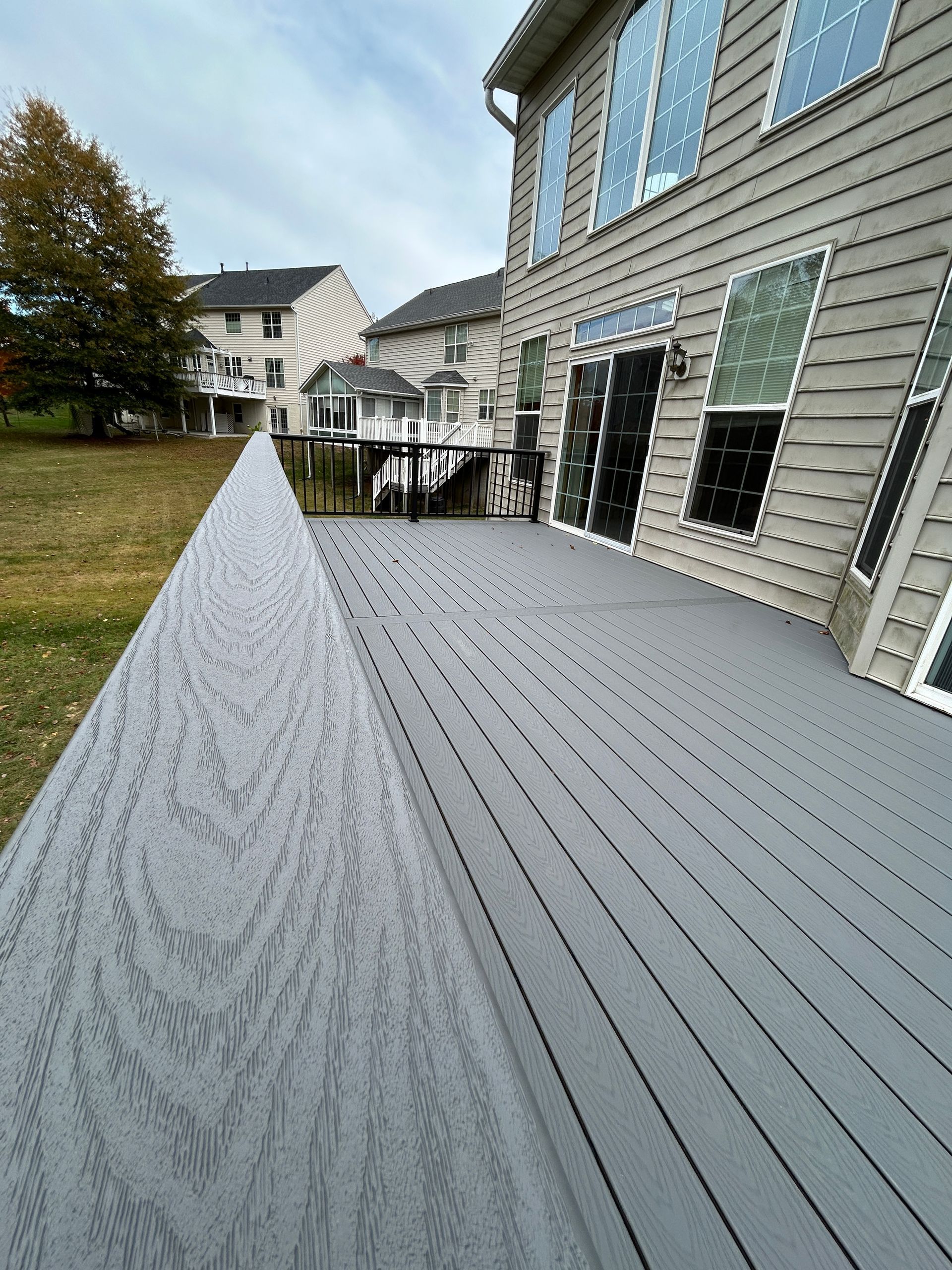 Gray composite deck with matching railing, adjacent to a beige house with several windows.