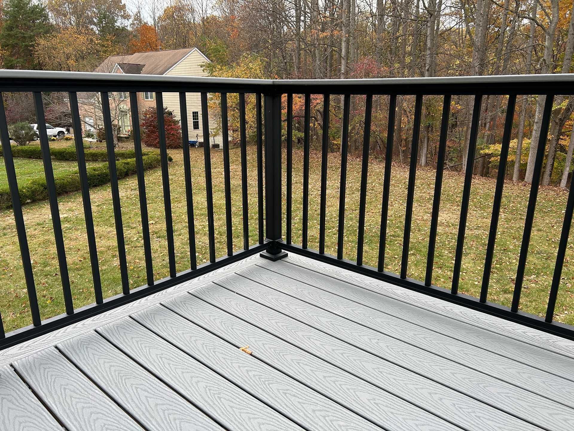 Corner of a deck with gray composite decking and black metal railing, overlooking a yard with trees.
