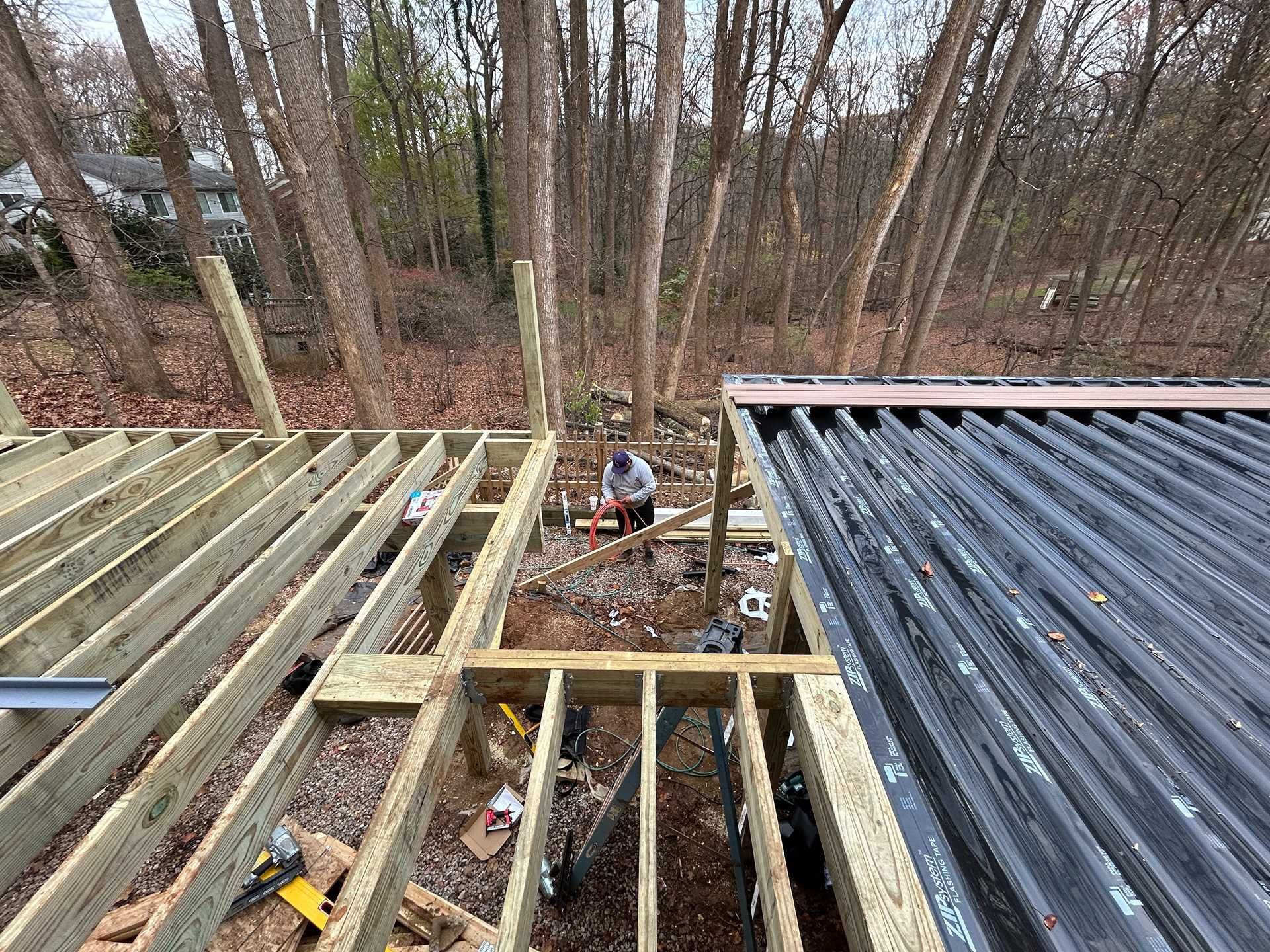 Construction site, deck framing. A person works amid exposed wooden joists; a new deck is under construction.