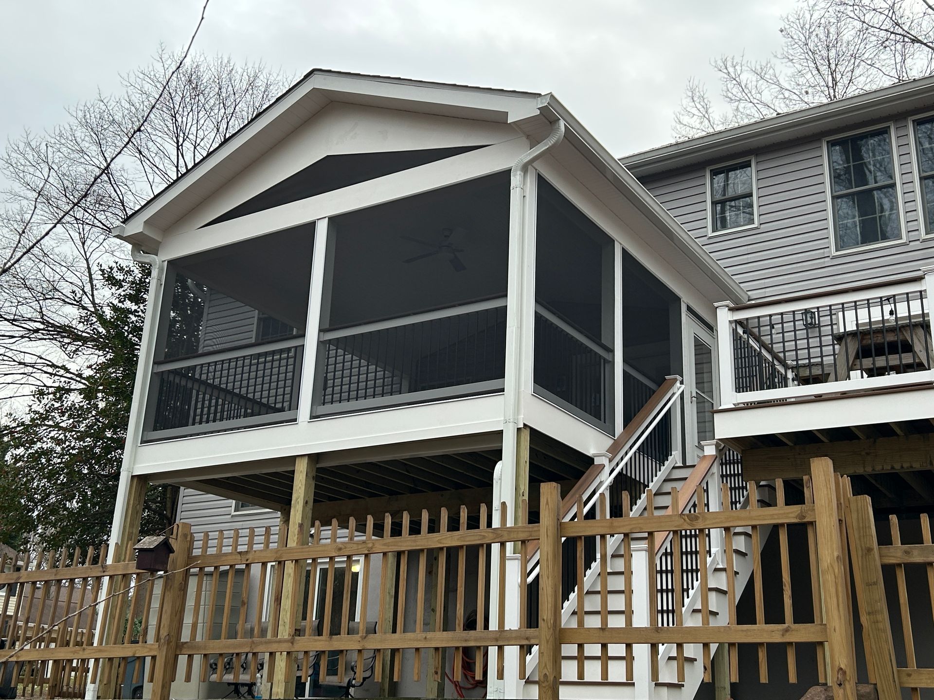 Screened porch addition on a raised deck, light-colored with dark screens, connected to a gray house, and a wooden fence.