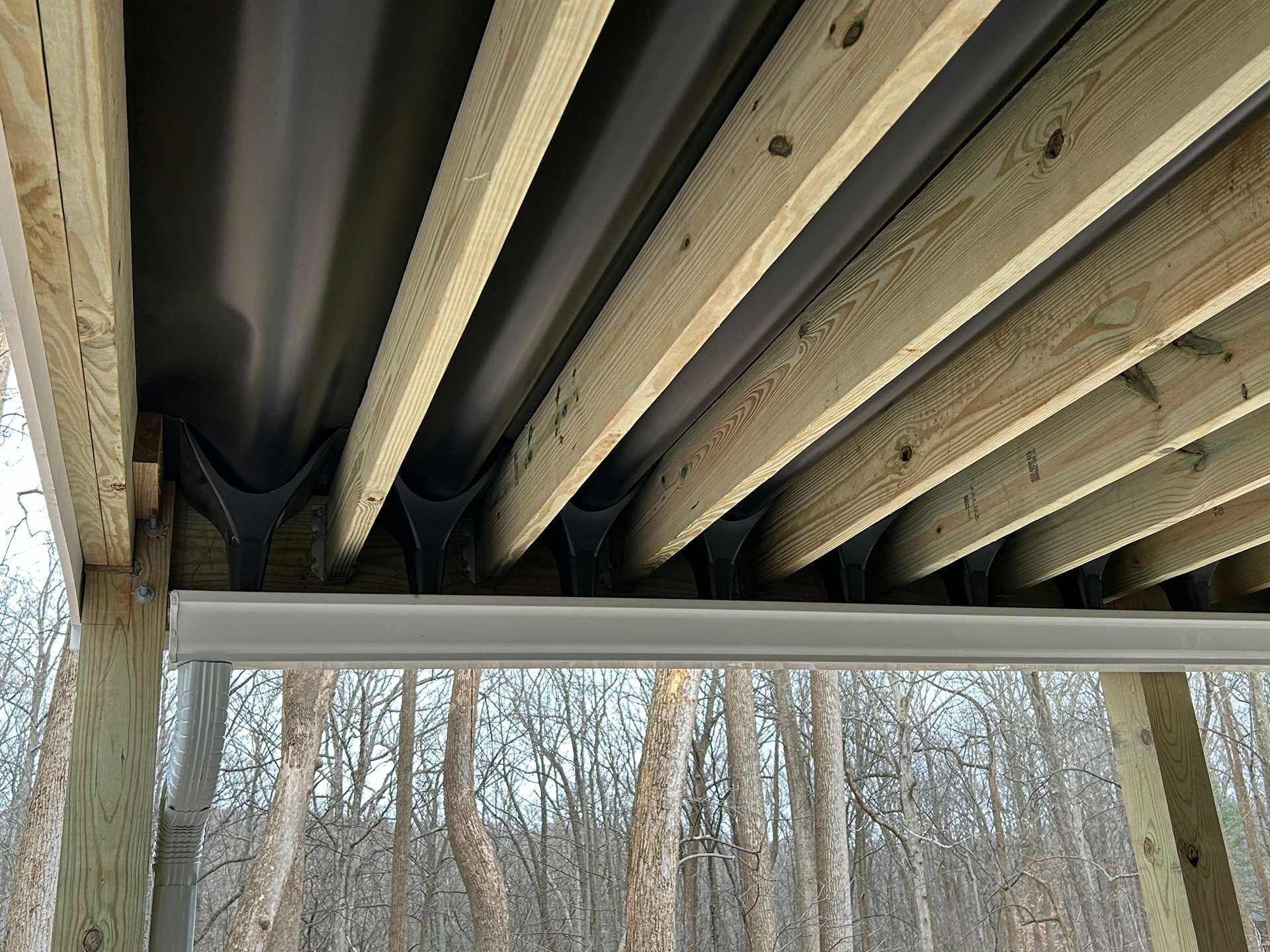 Underside of a deck showing wooden beams, dark corrugated metal, and a light-colored metal trim.
