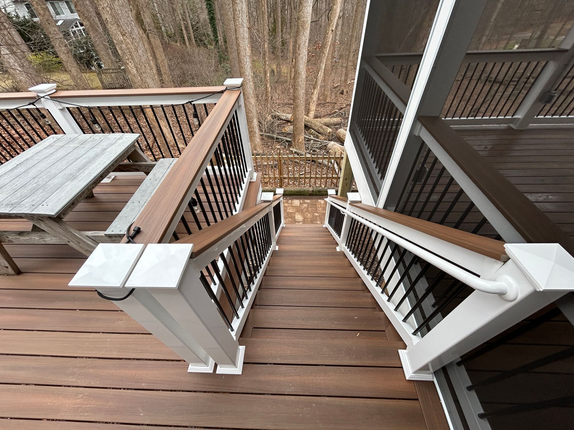 Wooden deck with stairs leading down into a wooded area, brown boards, white railing posts, and brown handrails.