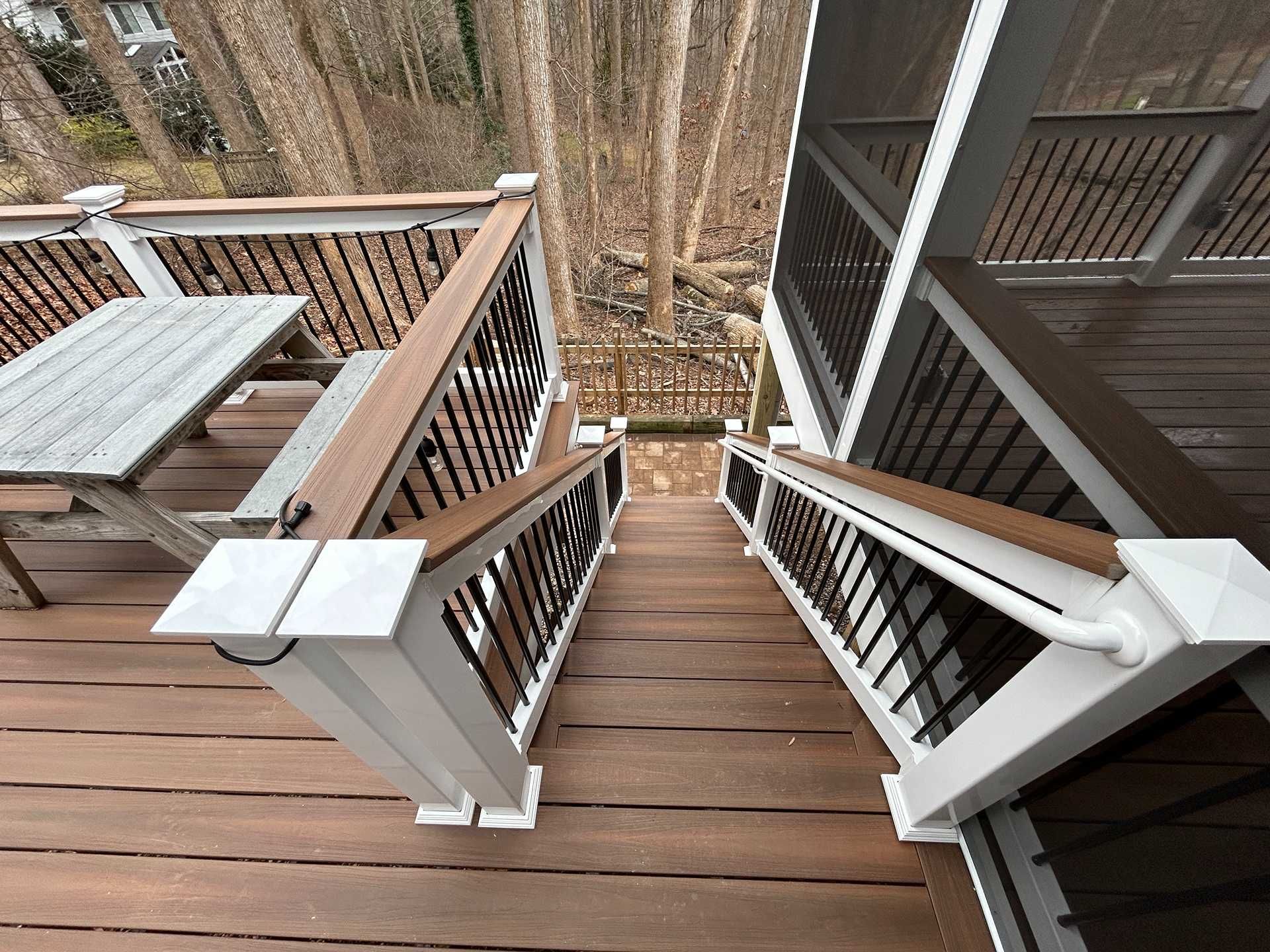 Wooden deck with stairs leading down to a wooded area. Black metal railings and a picnic table are visible.