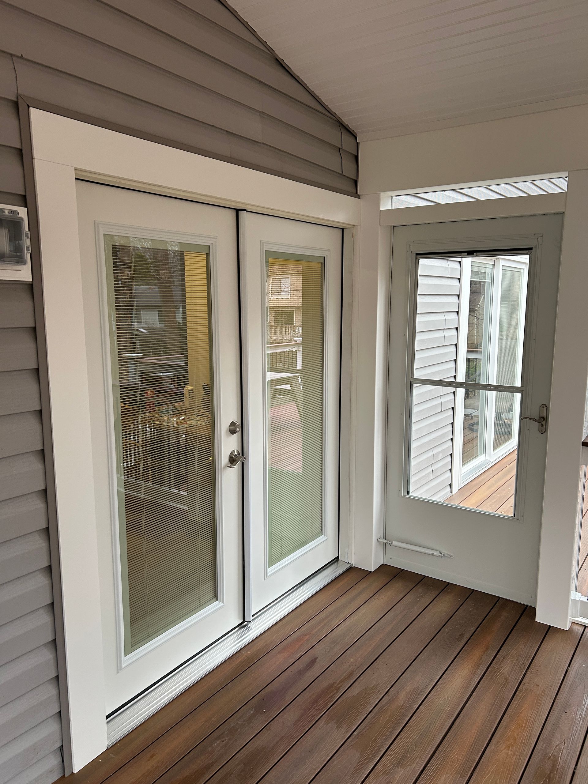 Double glass doors and a screen door on a wooden porch, all with white trim, siding.