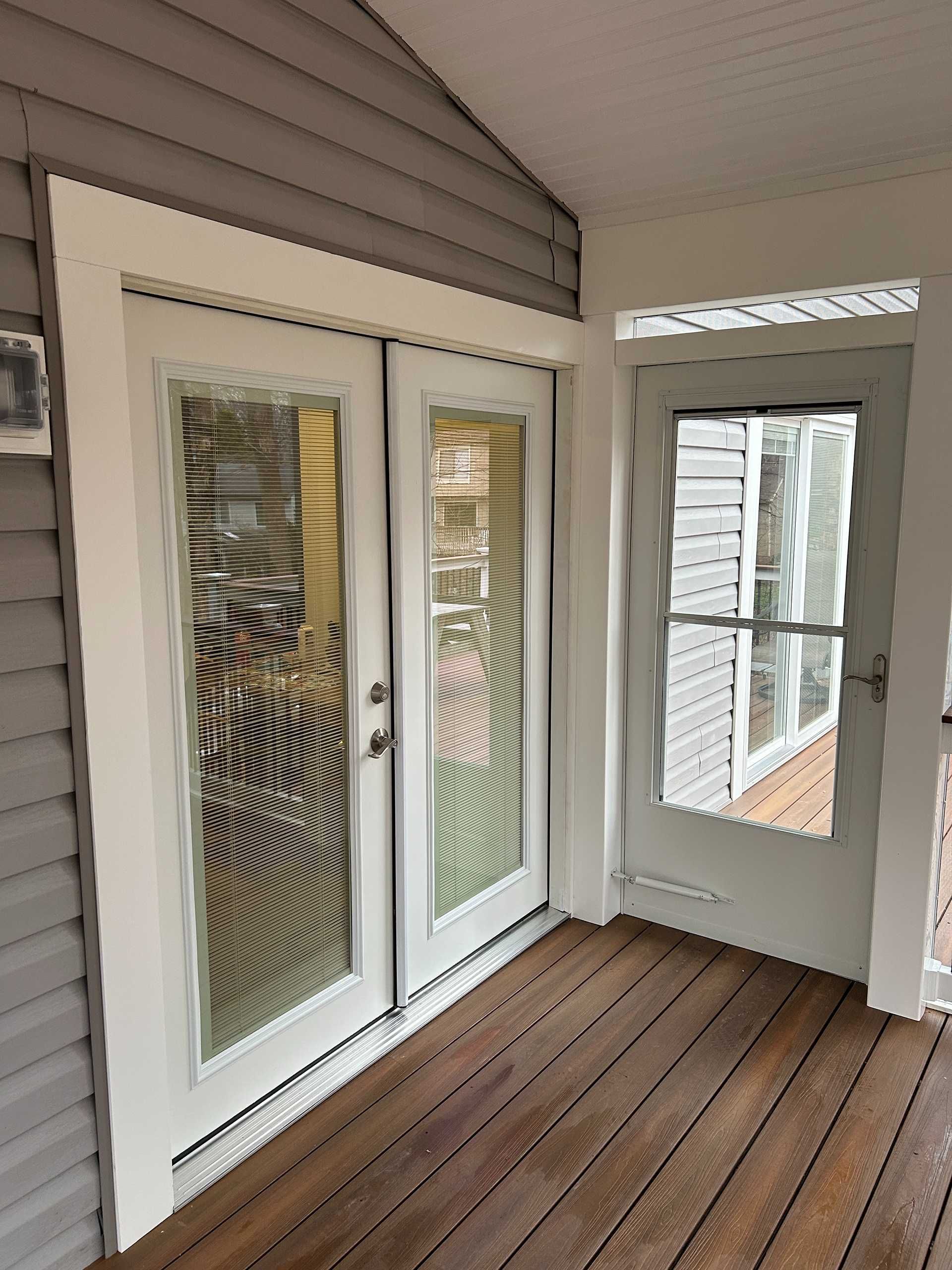 Two white French doors and a screen door on a wooden deck. The doors are framed with white trim and surrounded by gray siding.