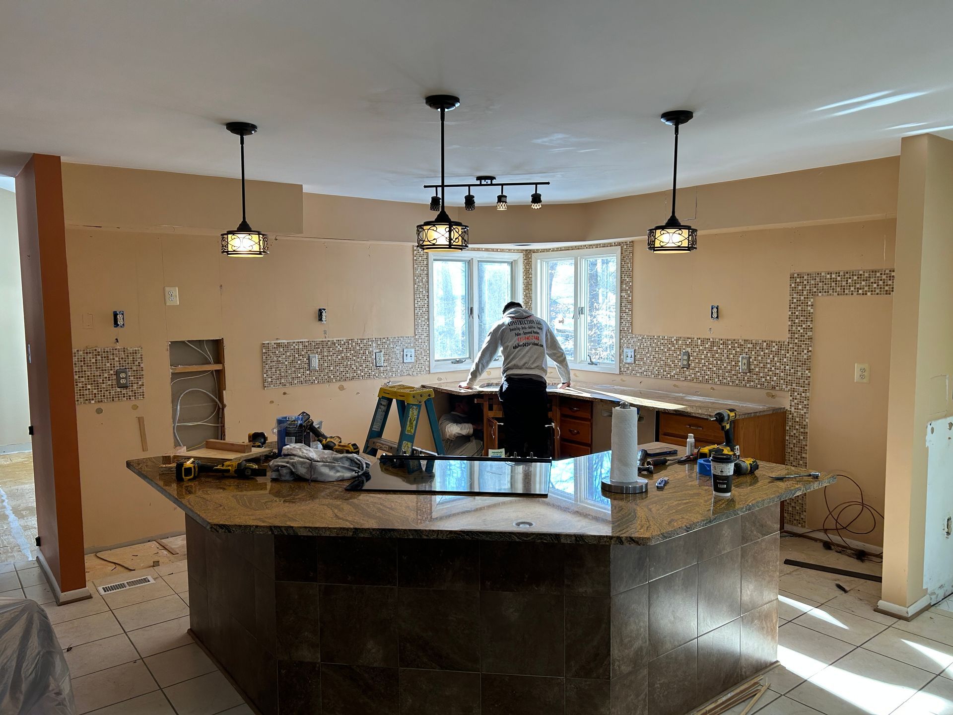 Kitchen undergoing renovation with island and a worker; neutral tan walls, pendant lights, and window.