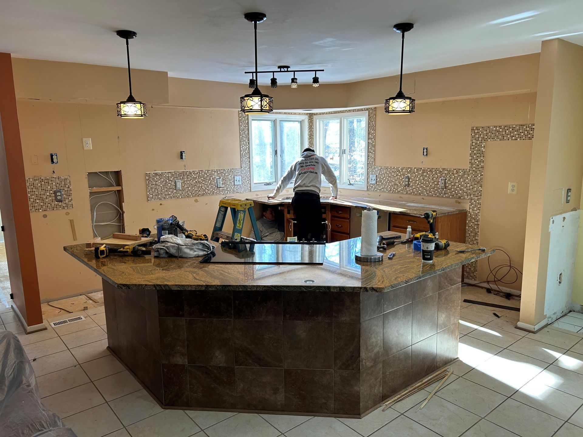 Kitchen under renovation with island and person working. Beige walls and tiled floor.
