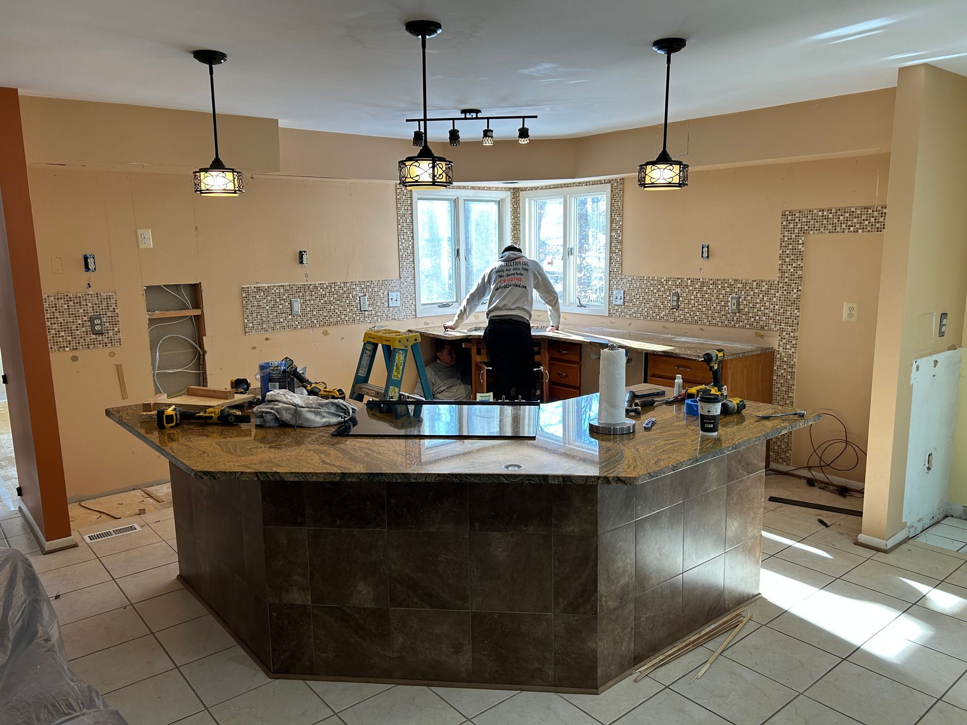 Kitchen undergoing renovation; person near window, island with granite countertop. Tiles removed from walls, tools visible.