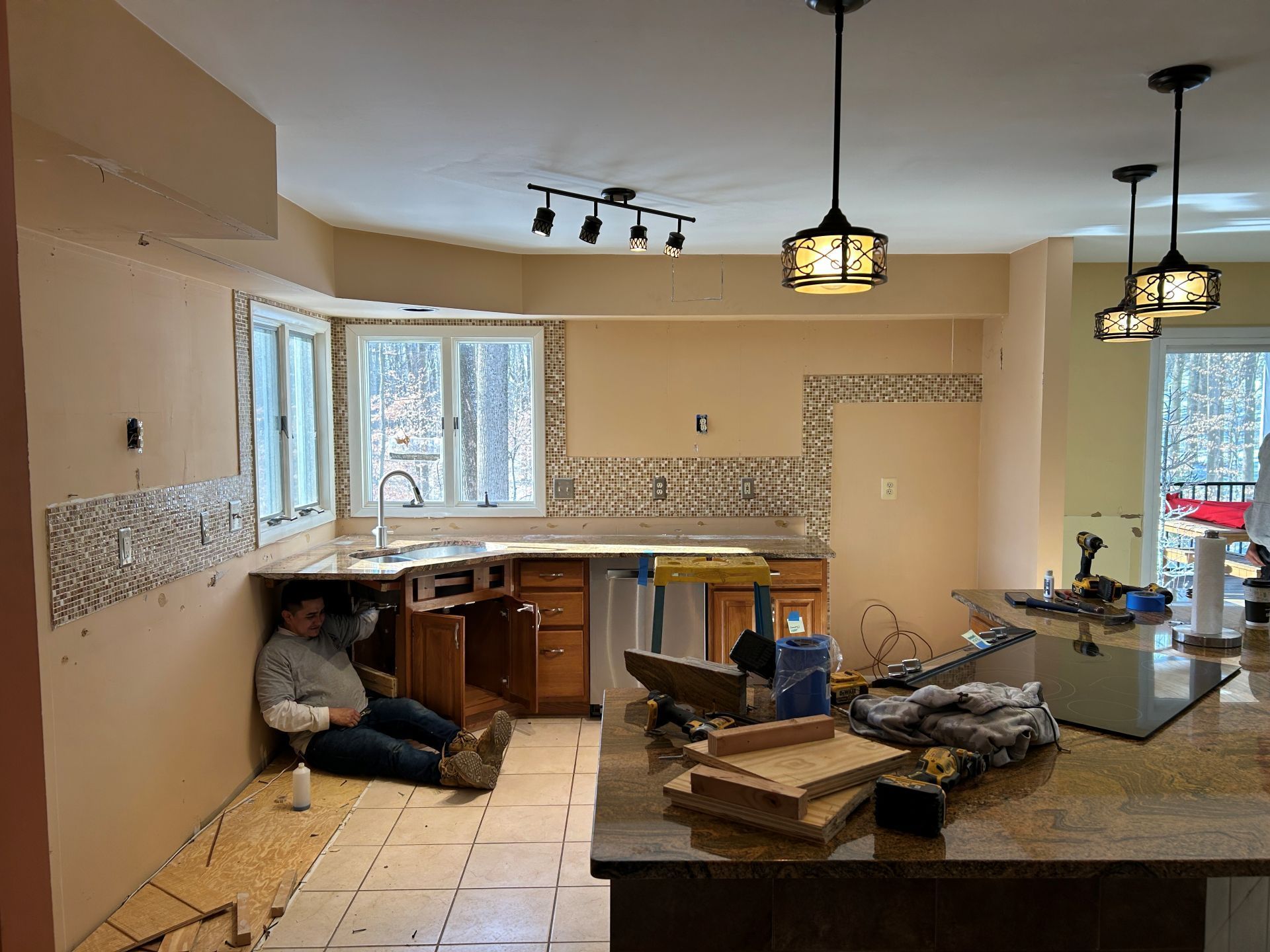 Kitchen under renovation, with worker resting under a counter. Countertops, cabinets, and tiling are visible.