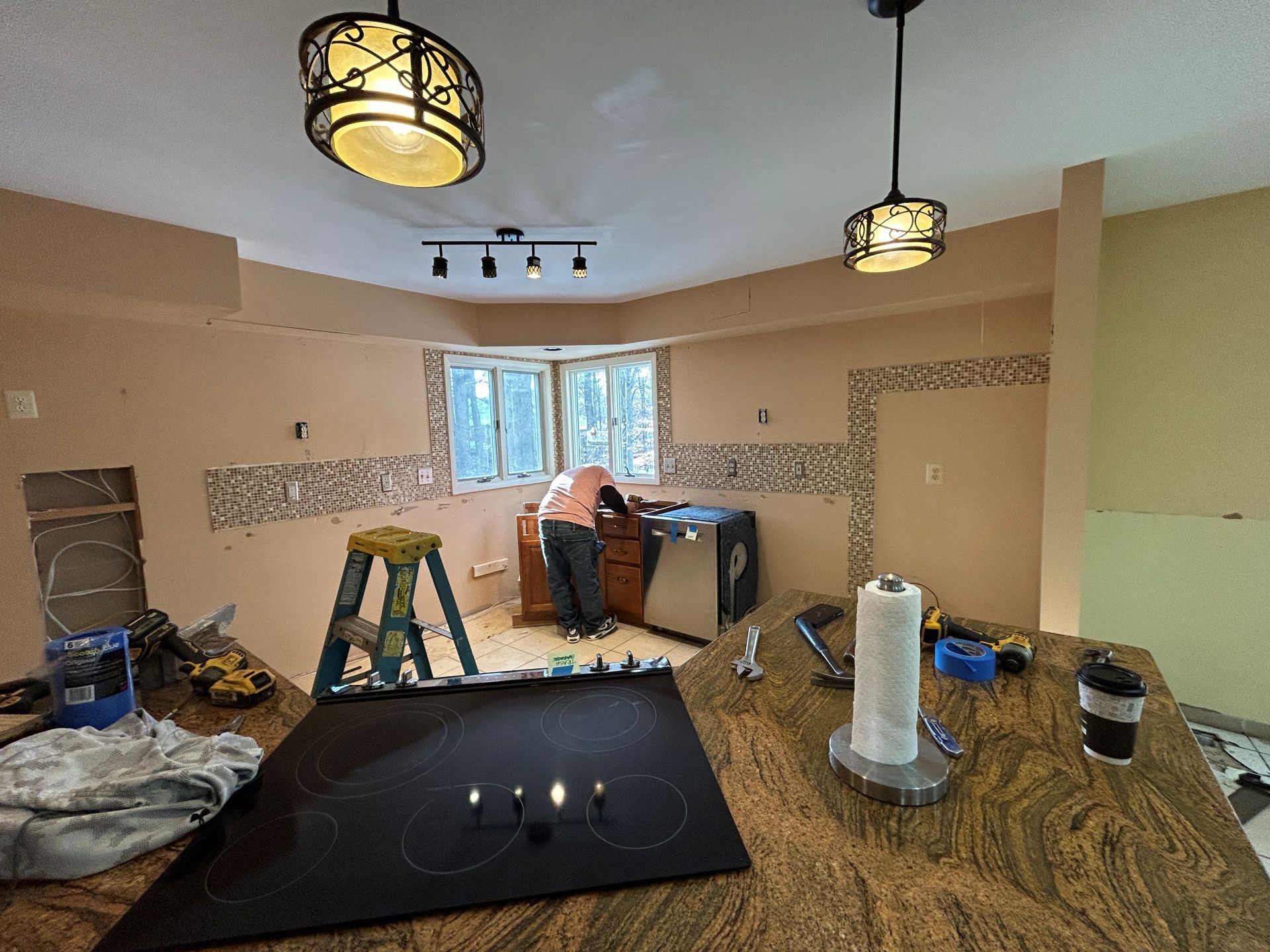 Kitchen renovation in progress, person working on cabinets. Tan walls, patterned tile, light fixtures, and a stovetop are visible.