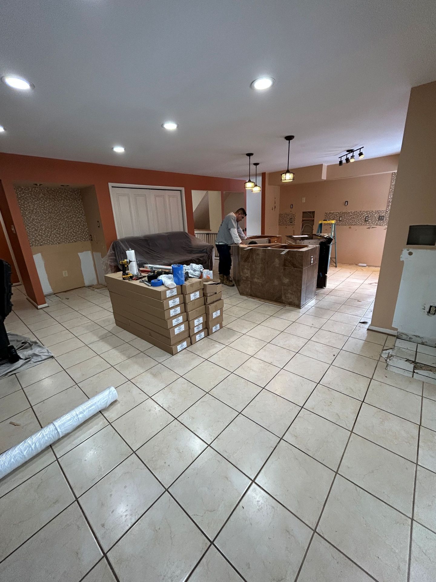 Kitchen under renovation, tile floor. Man near island, boxes, and partially removed cabinets. Neutral colors.