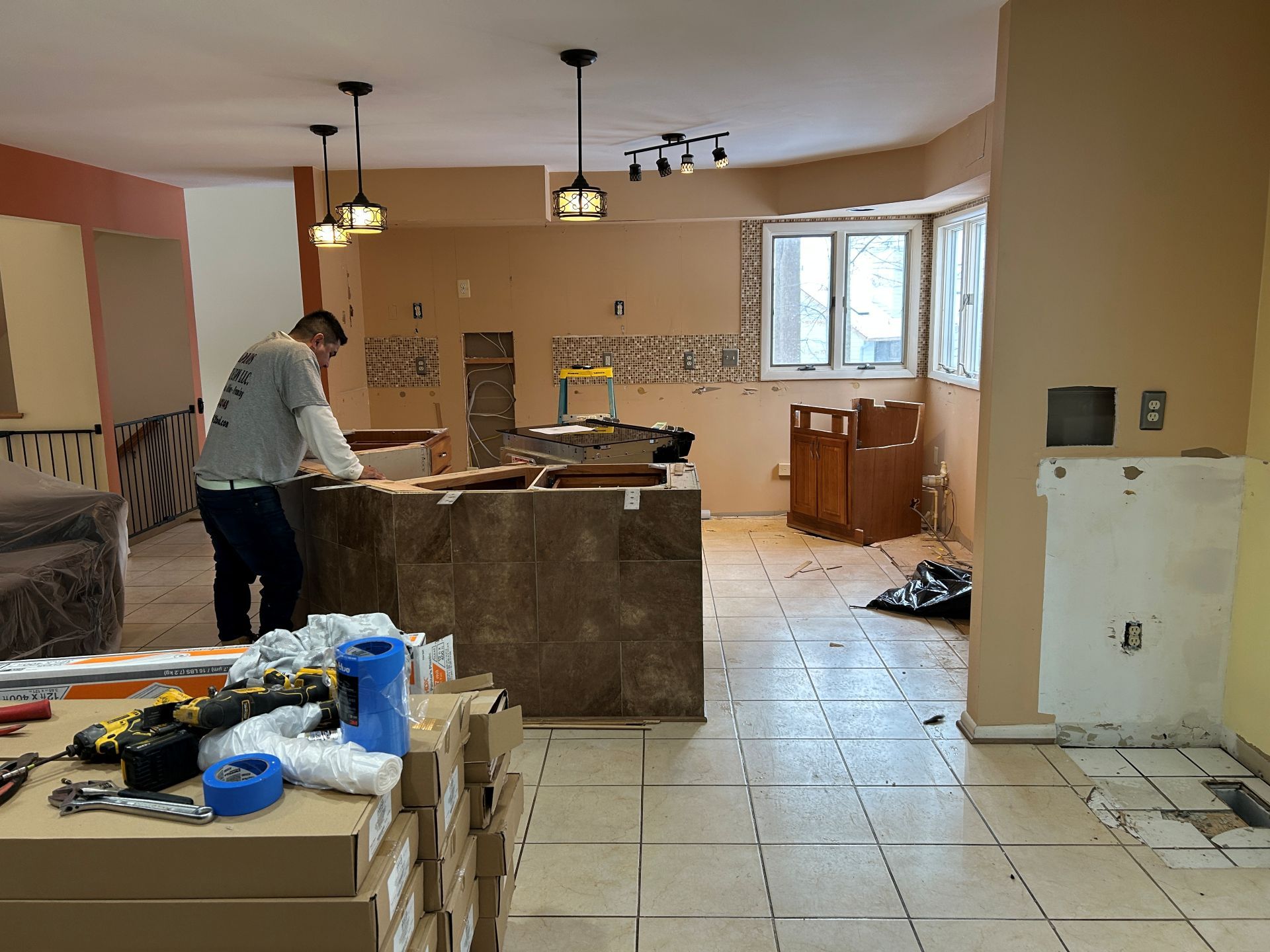 Man renovating a kitchen. New tile flooring installed, cabinets being worked on, boxes, tools. Beige walls.