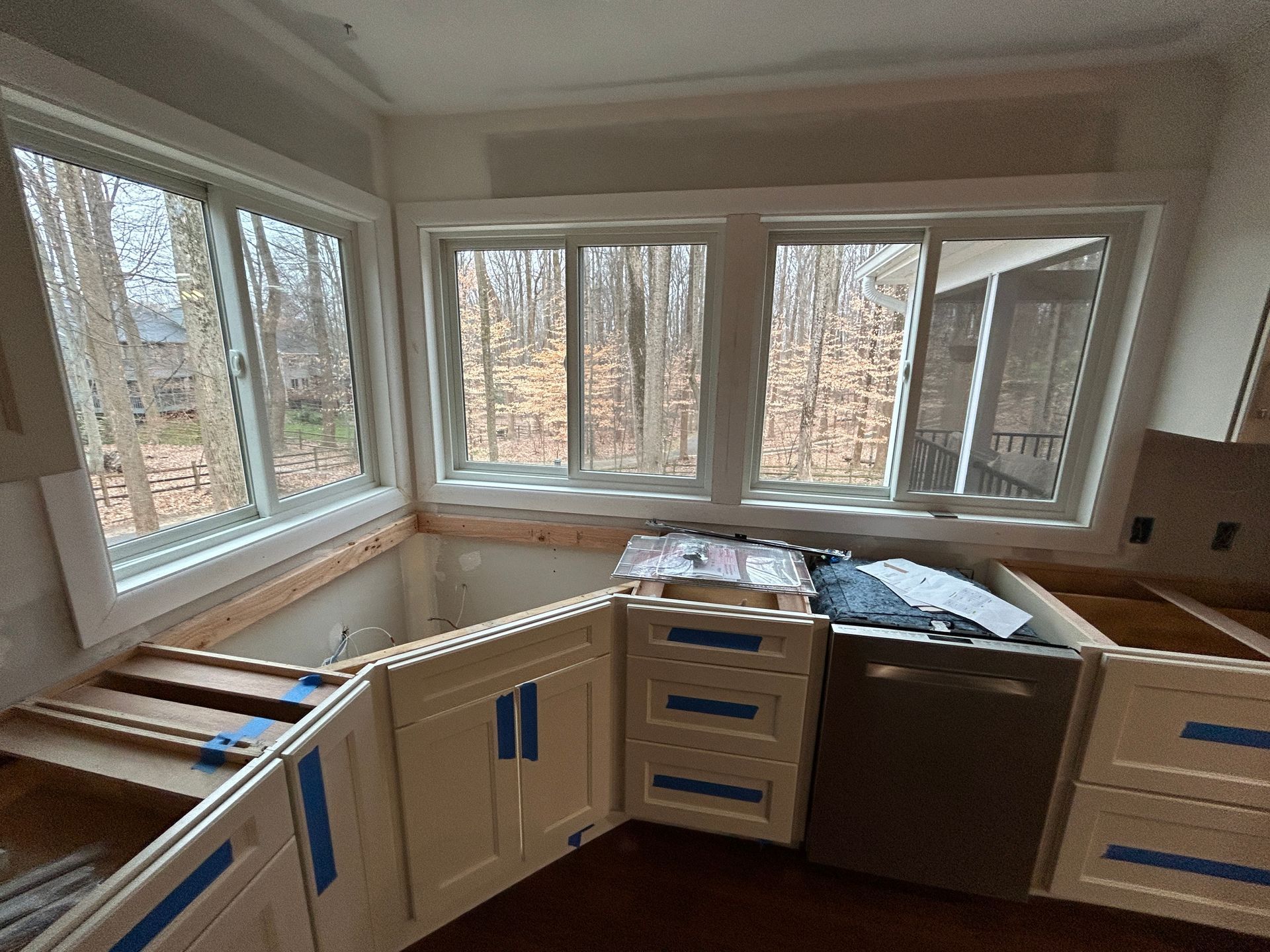 Kitchen cabinets being installed, with windows overlooking a wooded area. Unfinished cabinets and appliances are visible.