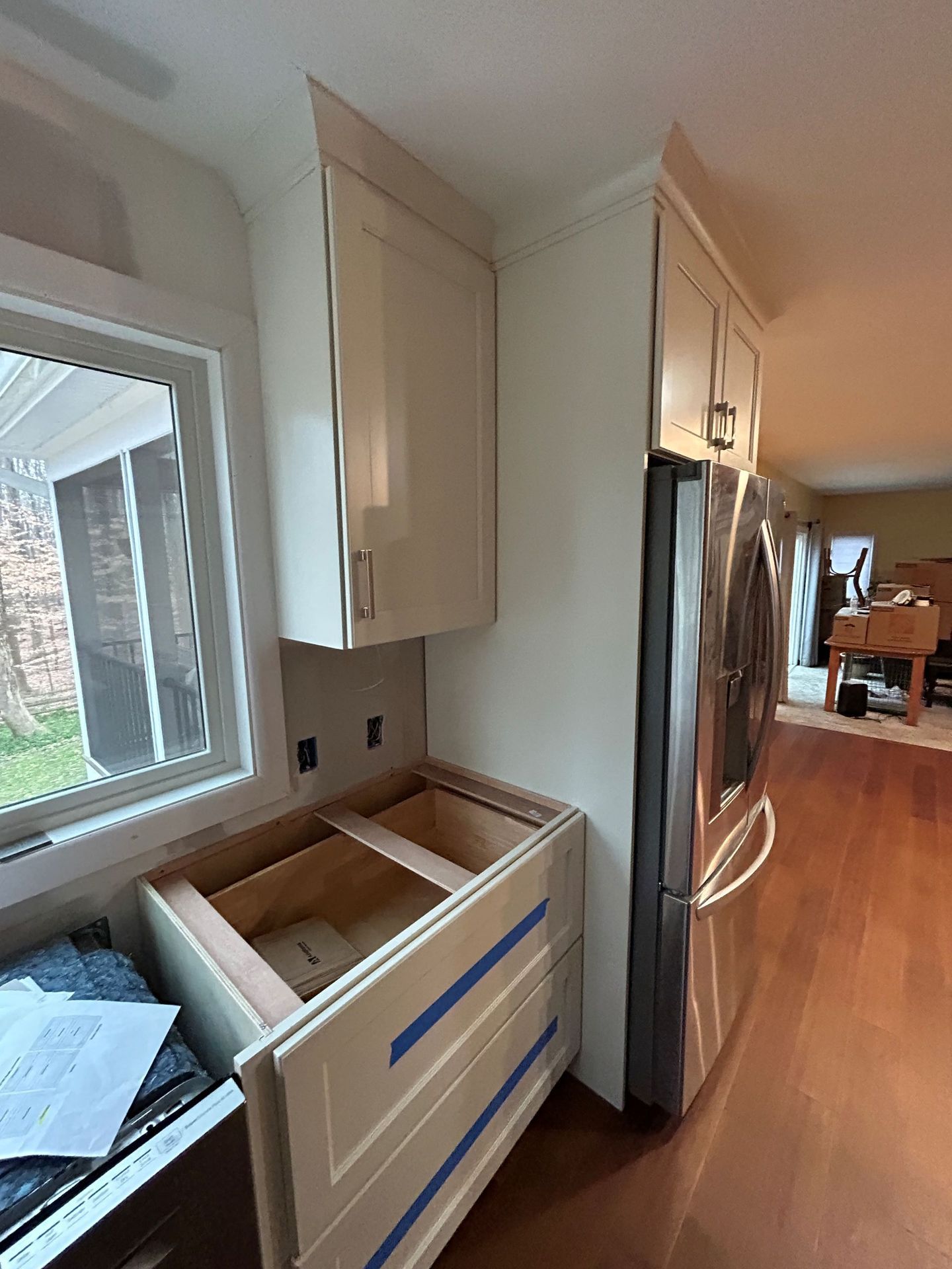 Kitchen cabinets under construction: cream cabinets, window, refrigerator, and unfinished lower cabinet.