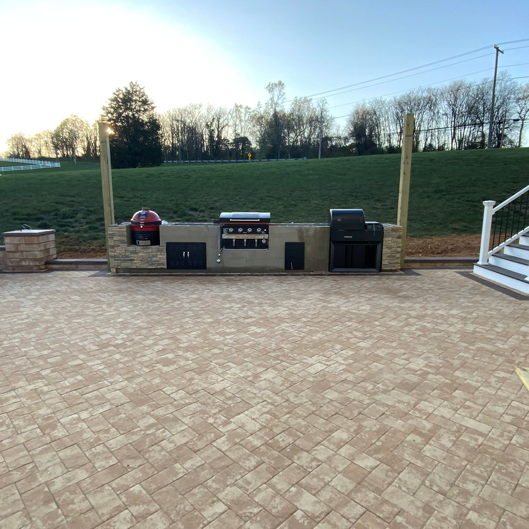Outdoor kitchen with grill, smoker, and storage on a brick patio with a grassy hill in the background.