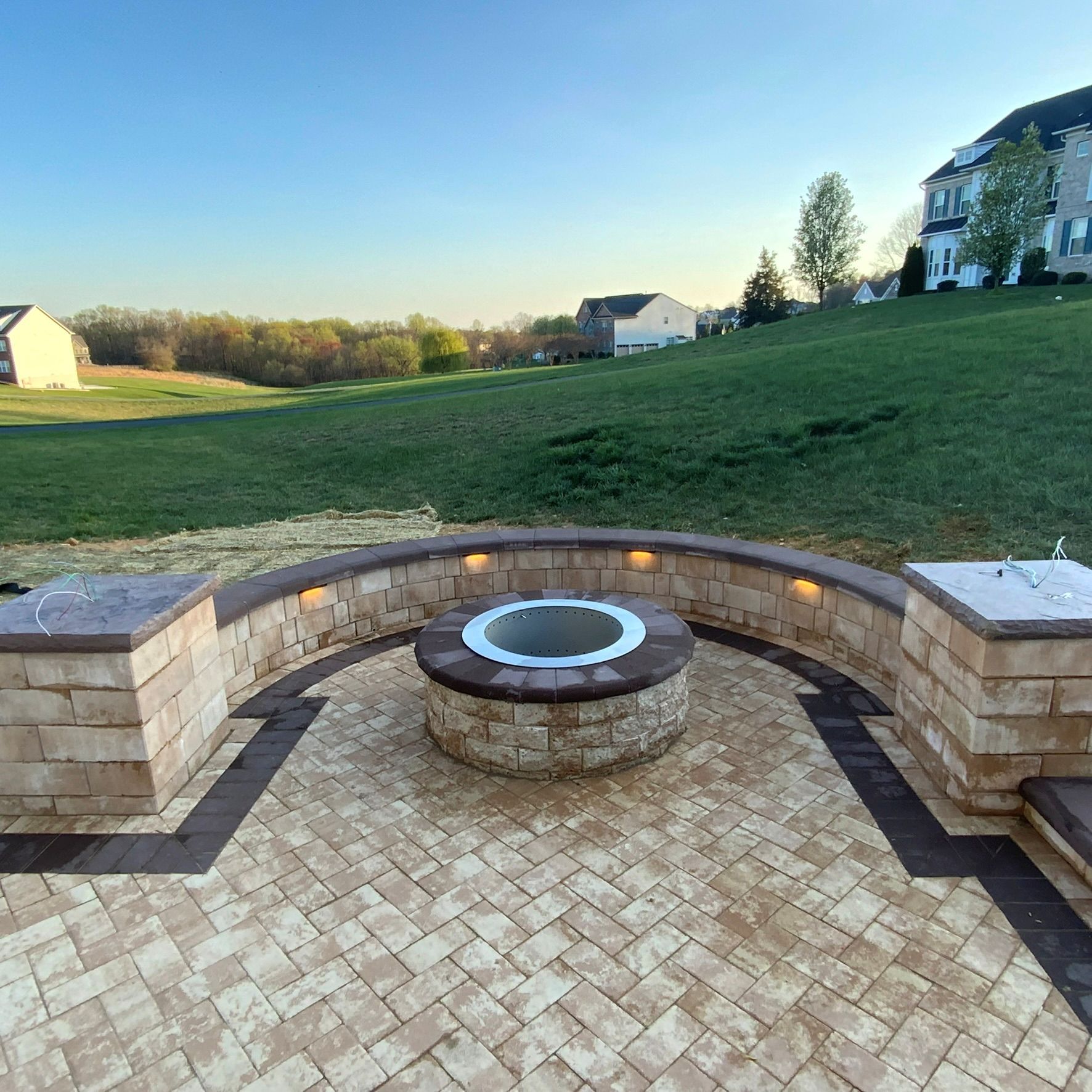 Brick patio with built-in fire pit and surrounding curved wall, overlooking a grassy hill, residential area in background.