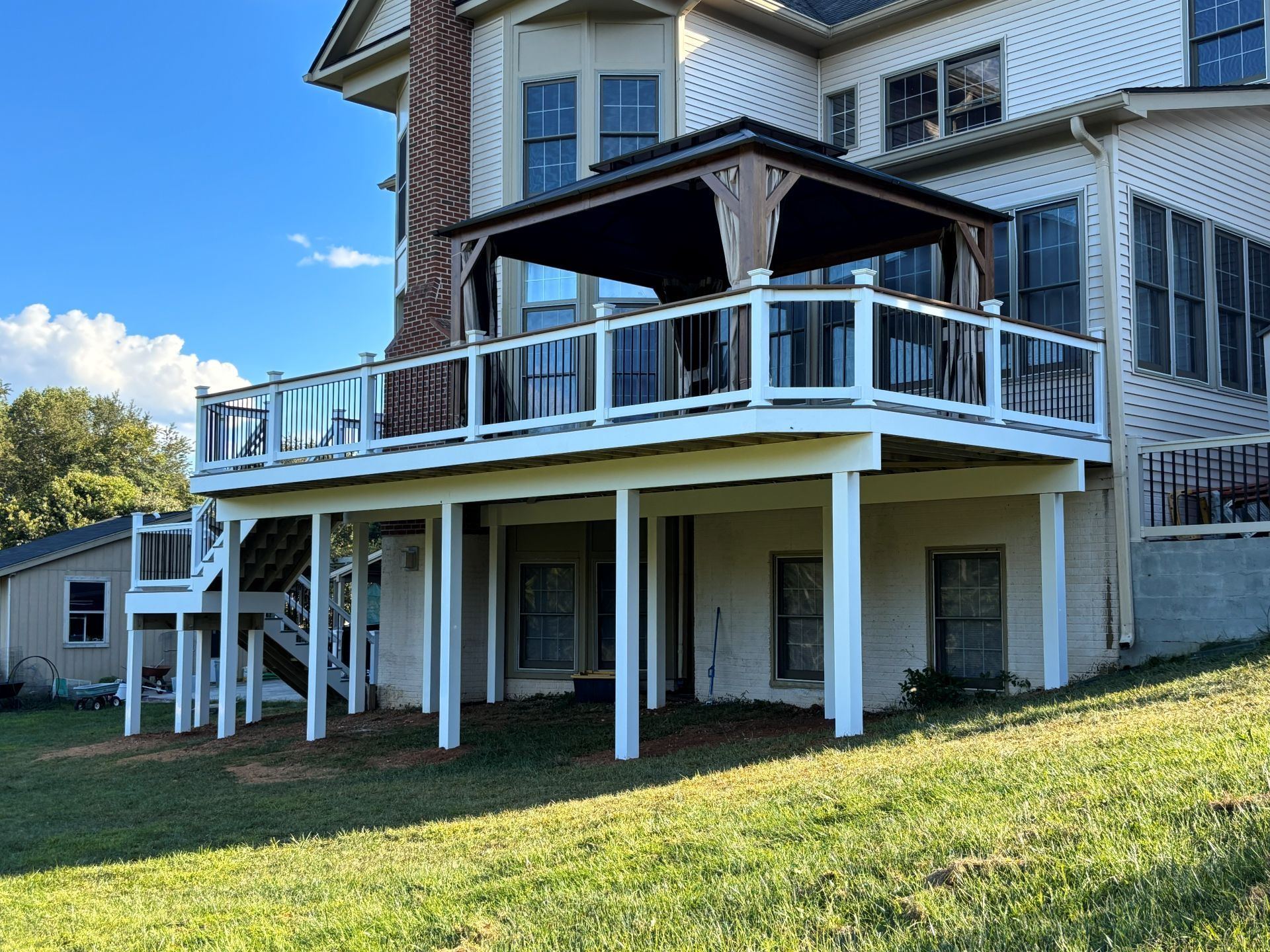 Two-story deck with a gazebo. White railing, glass panels, attached to a light-colored house. Green grass in foreground.