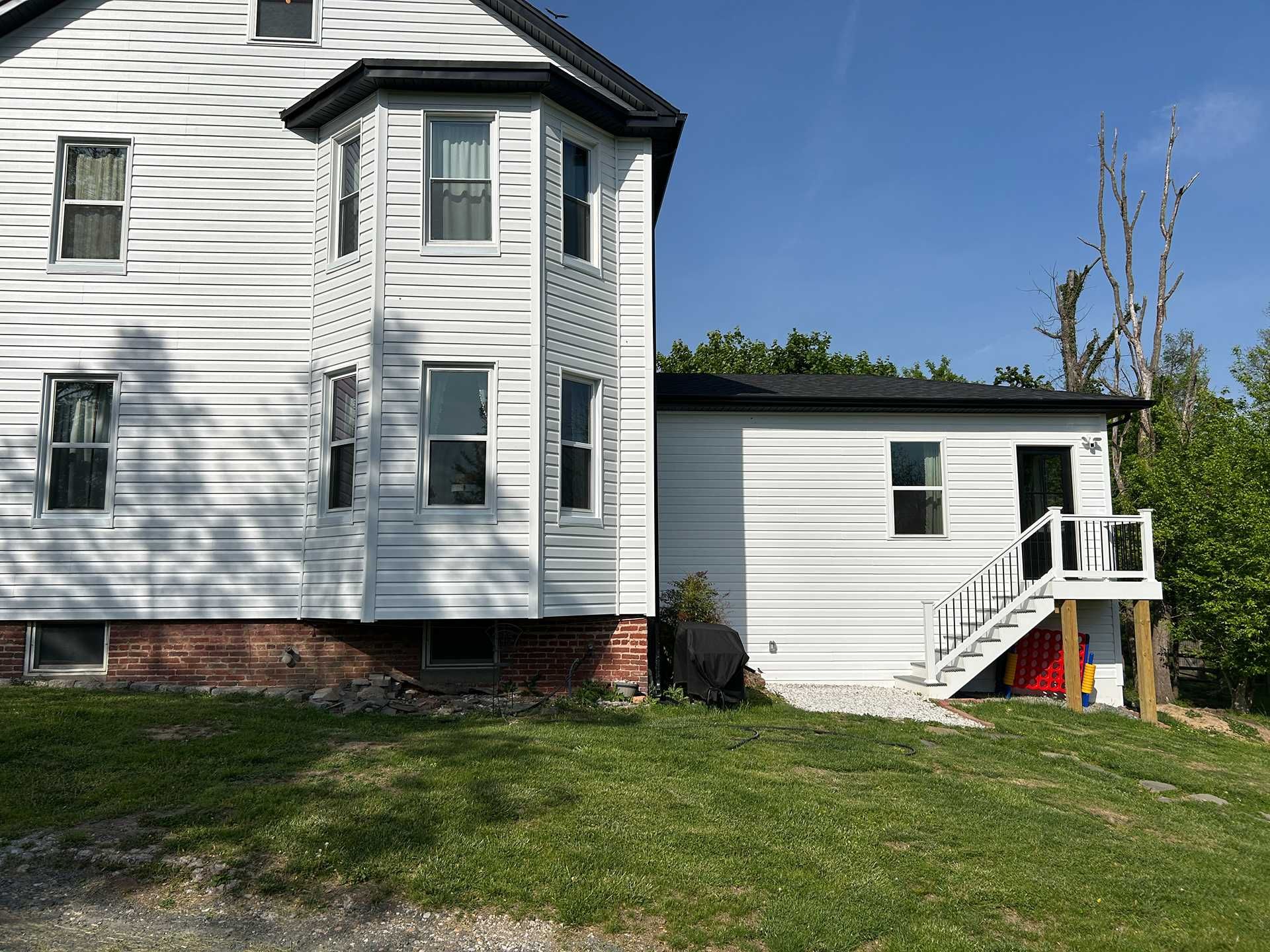 White house and attached structure with a bay window, steps, and door on a grassy hill under a blue sky.