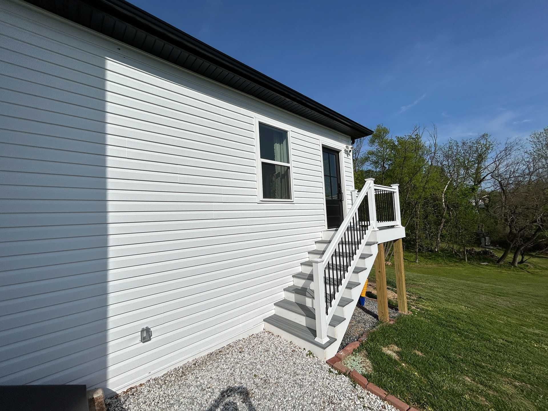 White house with stairs, a window, and a black door. Gray stone ground. Green grass and trees in background.