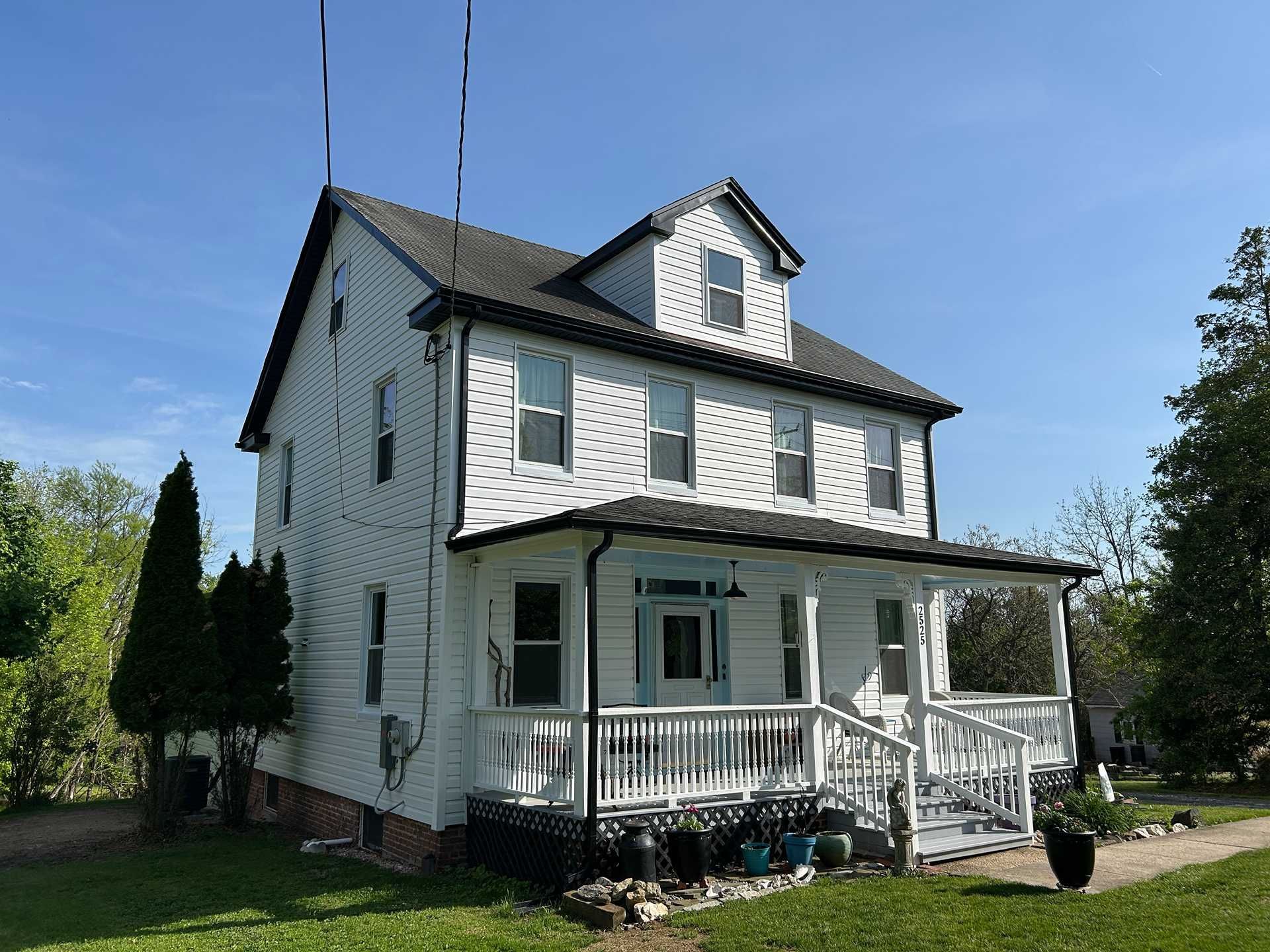 White two-story house with porch, black roof, and green lawn under a blue sky.