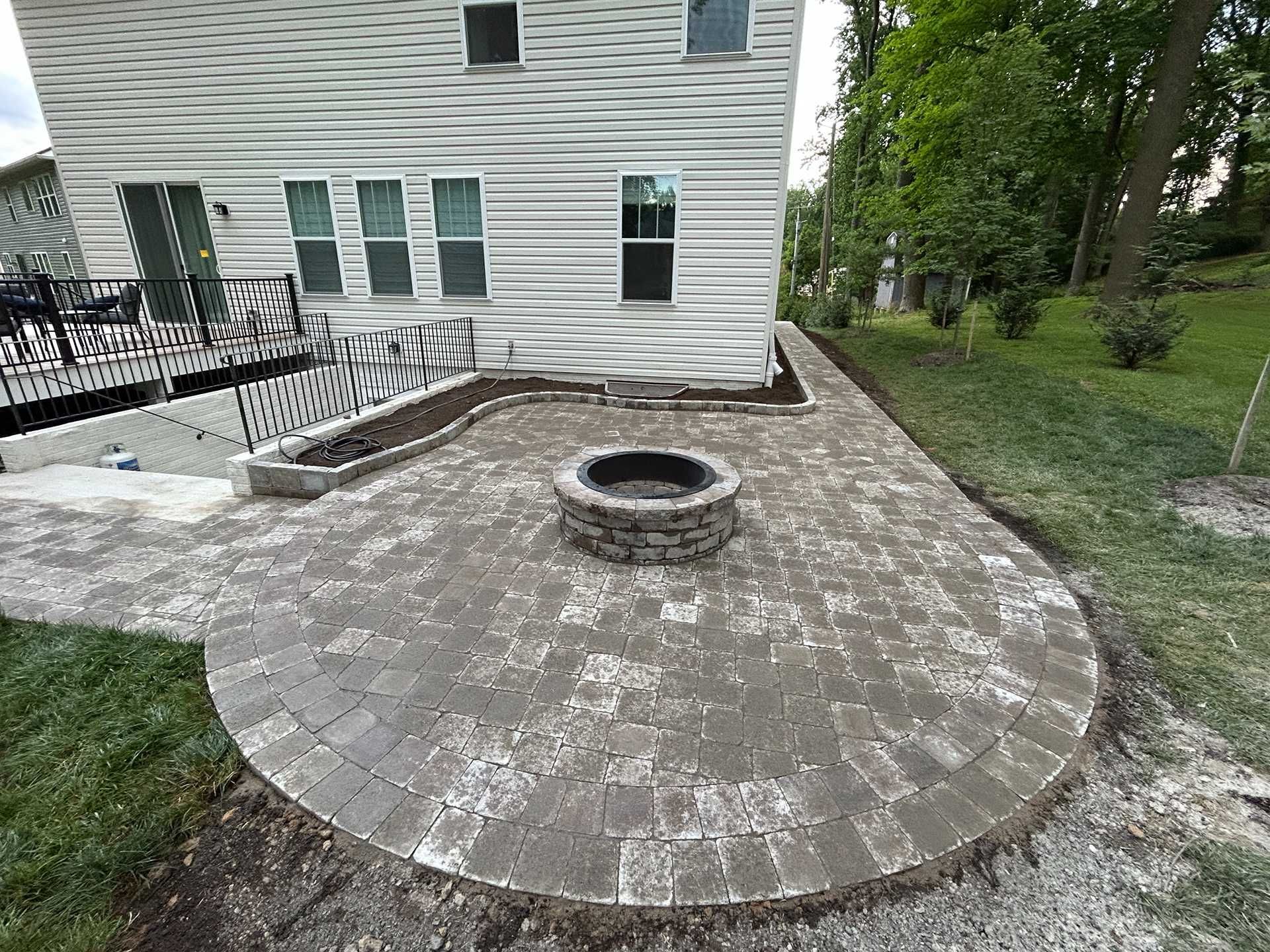 Stone paver patio with fire pit next to a house, edged with a grassy area.