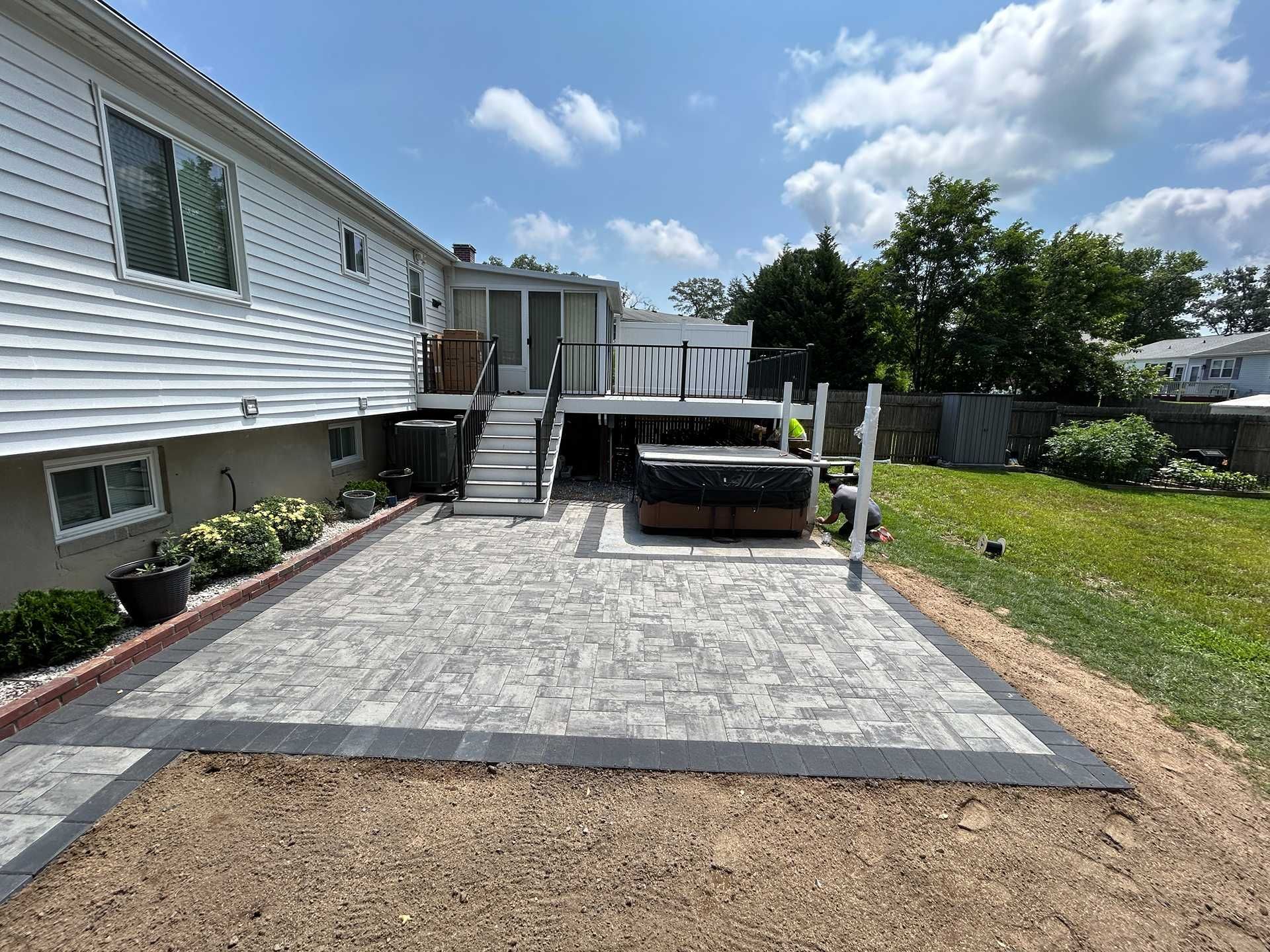 Backyard patio with hot tub, connected to a deck and house. Paved patio area with bordering gravel.