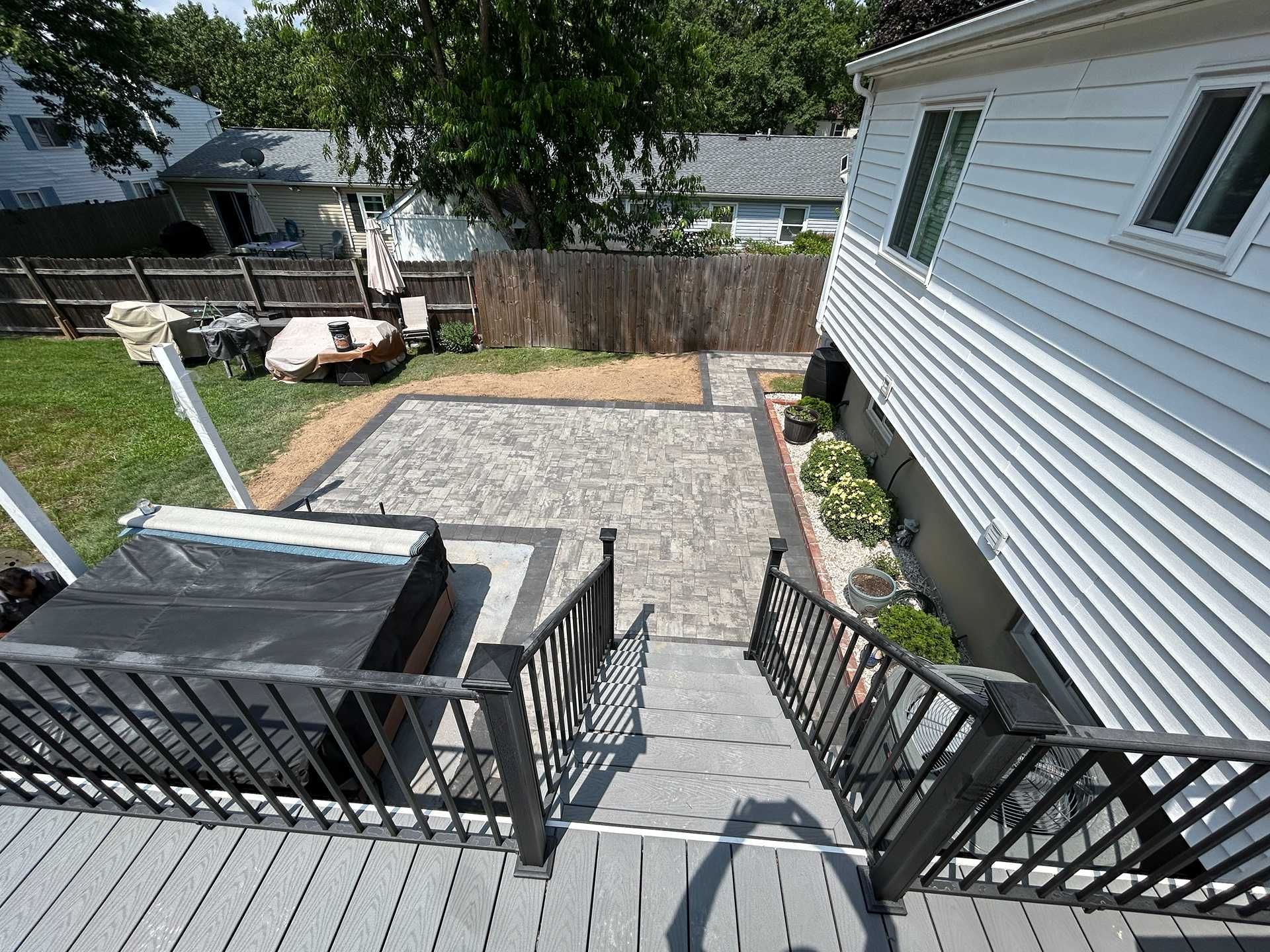 Deck stairs lead to a patio with a hot tub, bordered by landscaping and a fence.