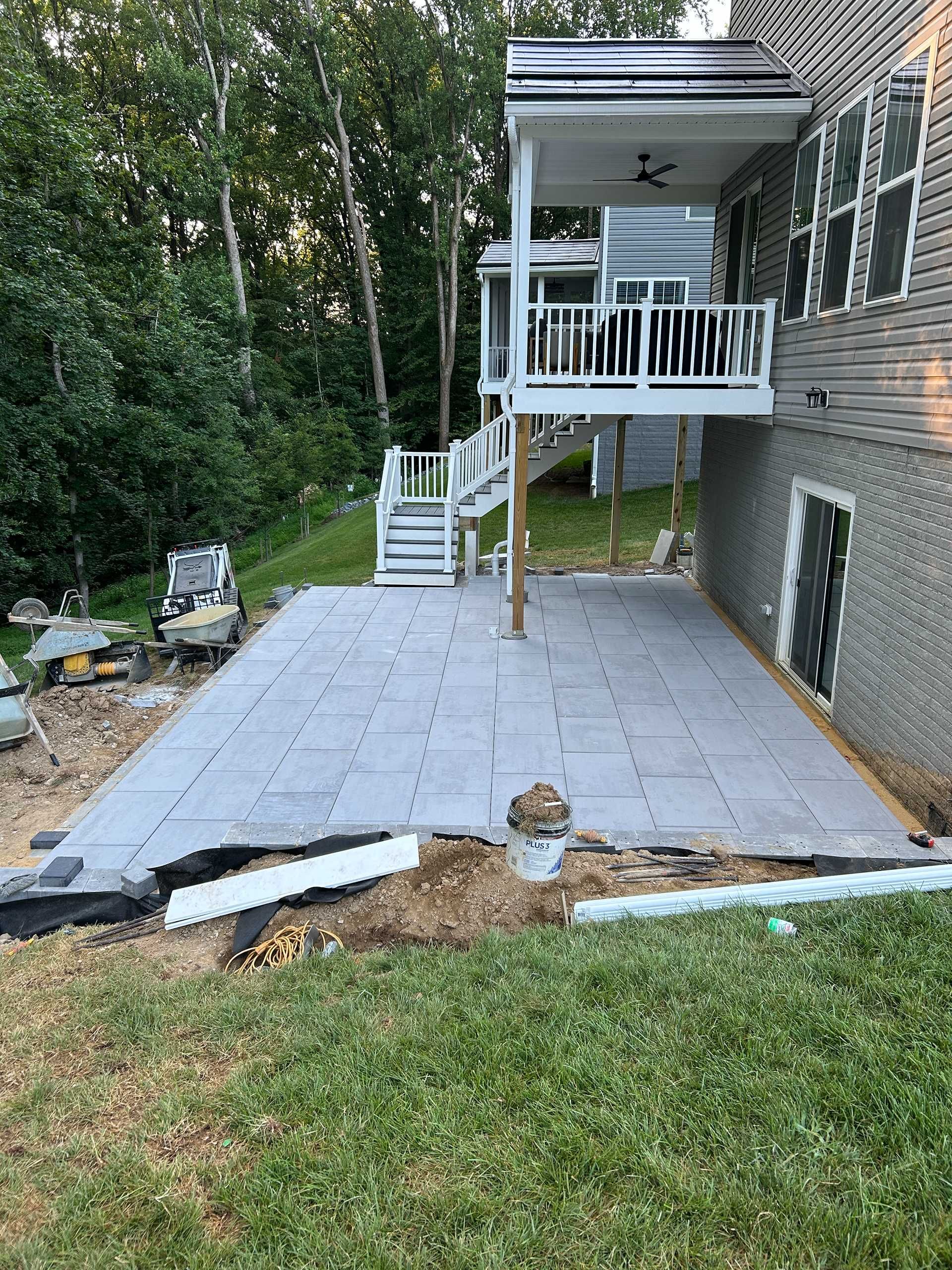 Patio construction: workers laying gray pavers on a leveled area next to a house with a raised deck and stairs.