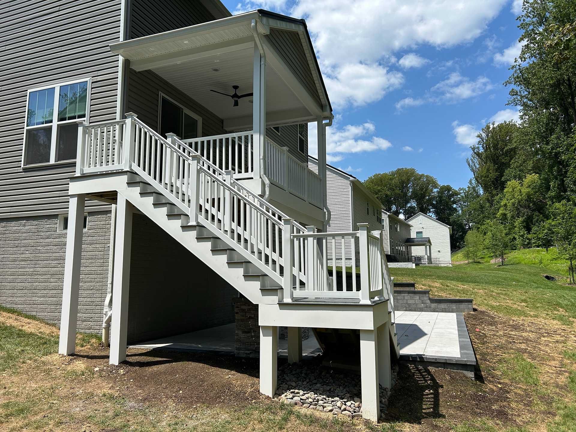 A two-level deck with stairs leads to a covered porch with a ceiling fan. Bright blue sky overhead.
