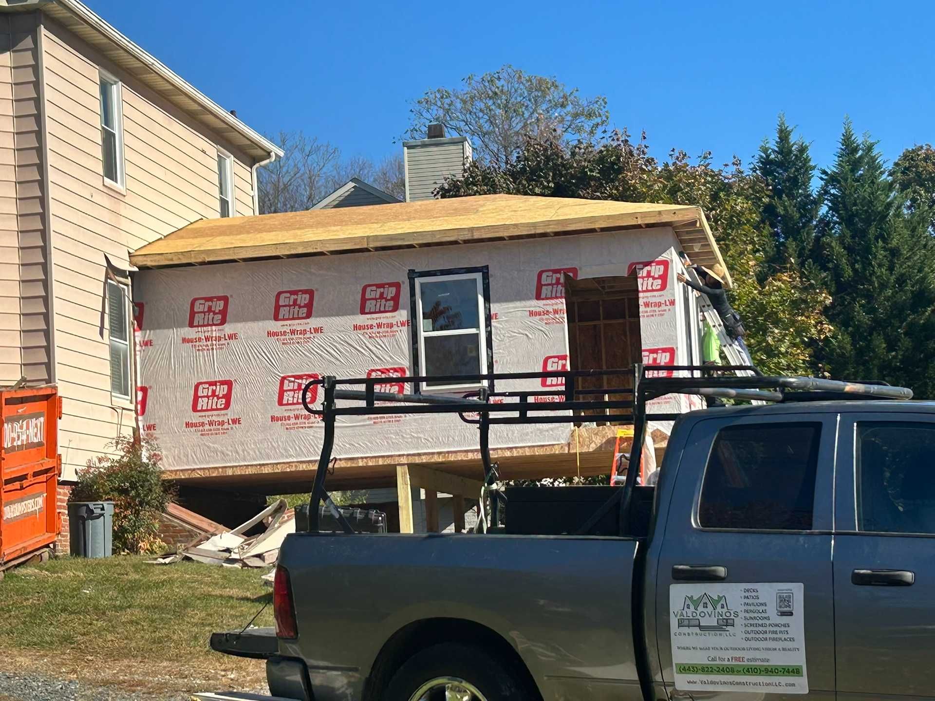 Construction of a small building addition next to a house; plywood roof and red-wrapped walls; a truck parked in front.