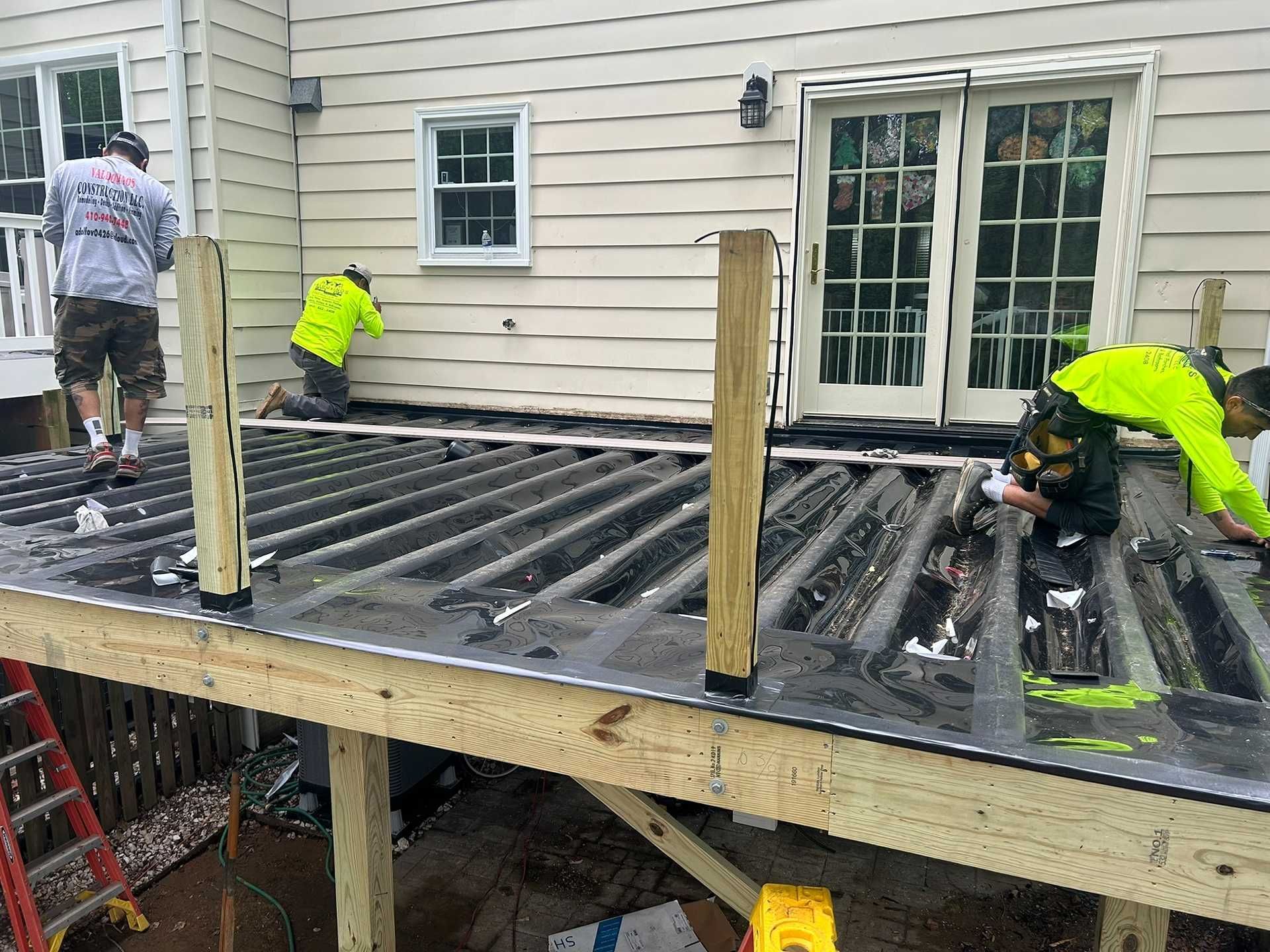 Workers installing decking on a wooden structure next to a house with sliding glass doors; a ladder is visible.