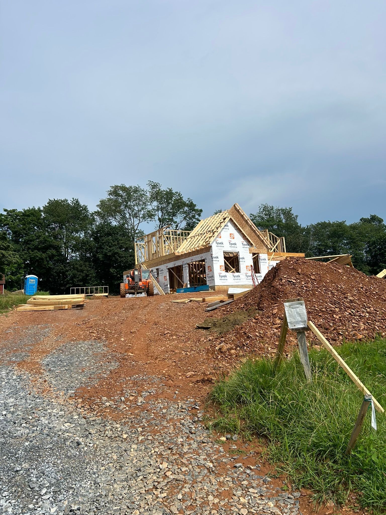 Construction site: house frame, wrapped in paper, roof partially complete; dirt pile in foreground.