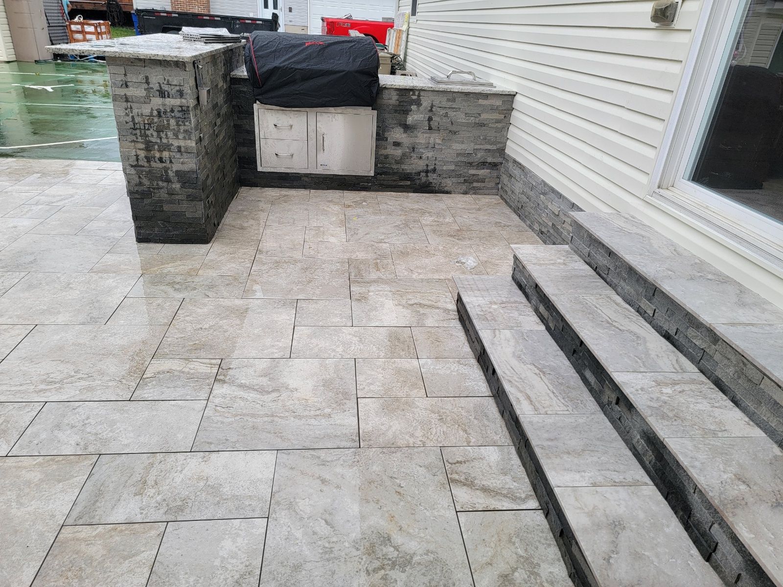 Outdoor patio with stone pavers, built-in grill area, and steps leading up to a house with white siding.