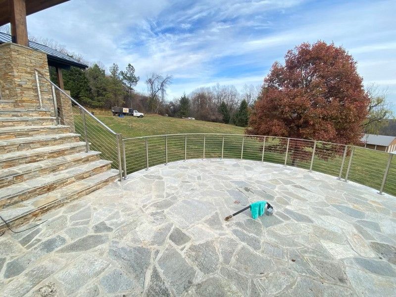 Stone patio with stainless steel cable railing, stairs, and grassy hillside. Bright blue sky.