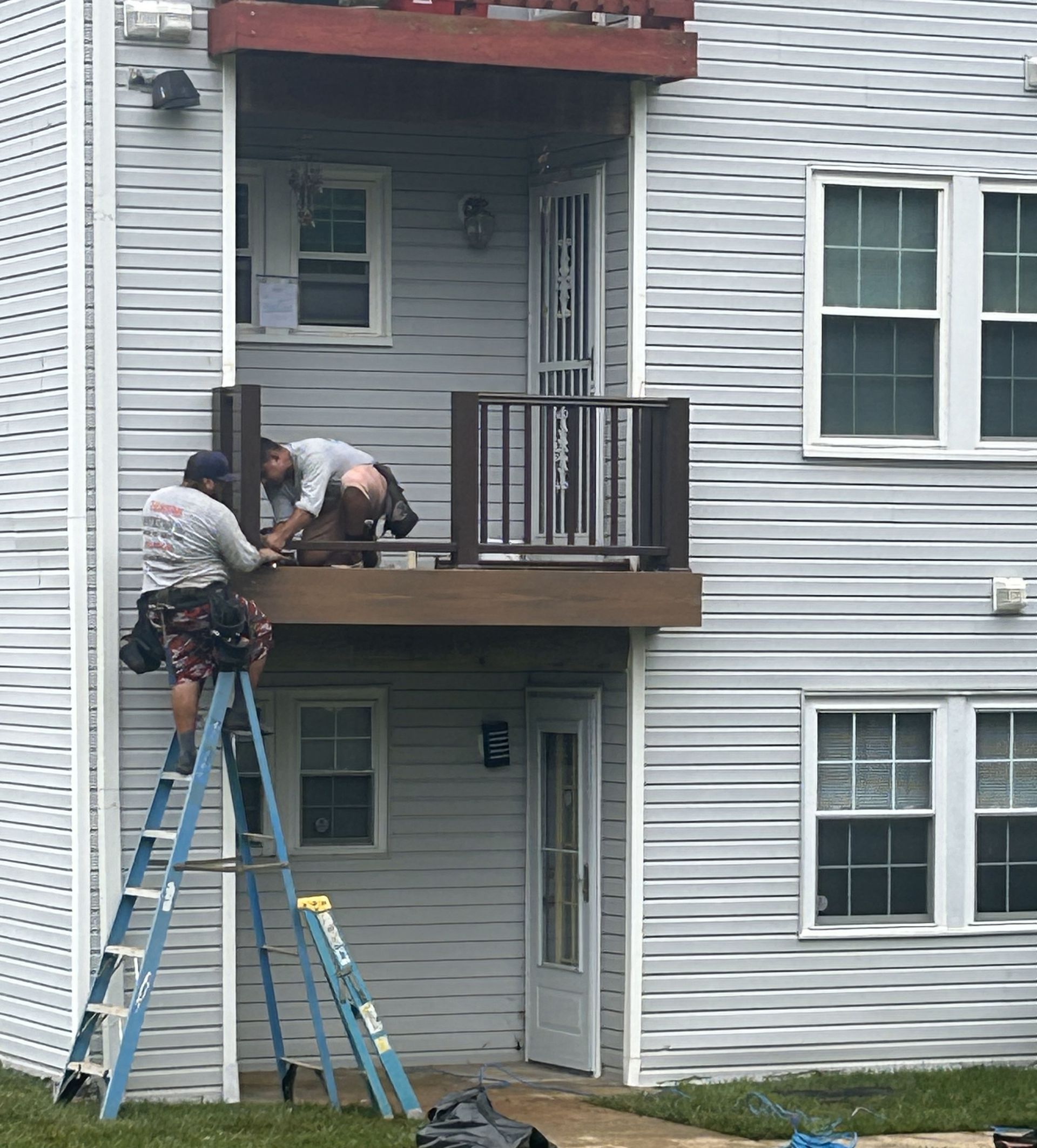 Two workers installing a deck on an apartment building. One on a ladder, one on the deck. Exterior shot.