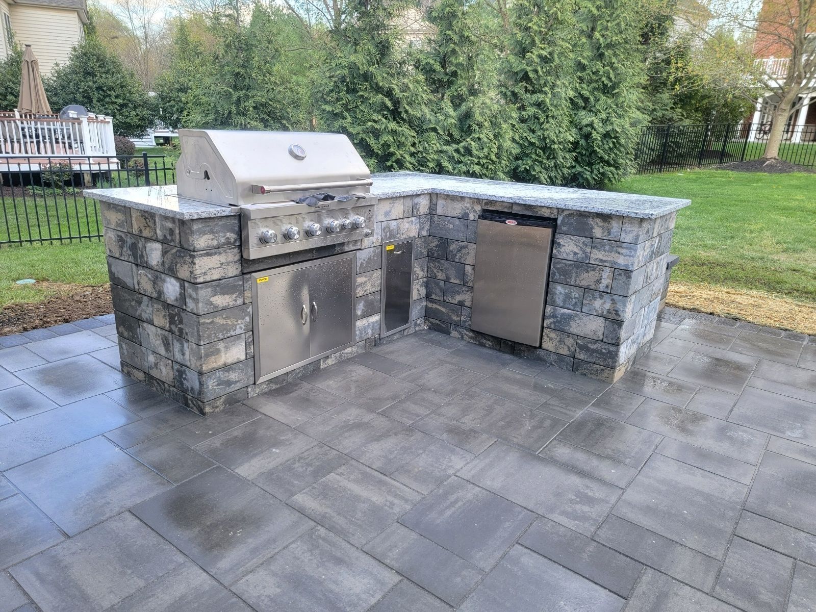 Outdoor kitchen with stainless steel grill, fridge, and stone facade on a paver patio.