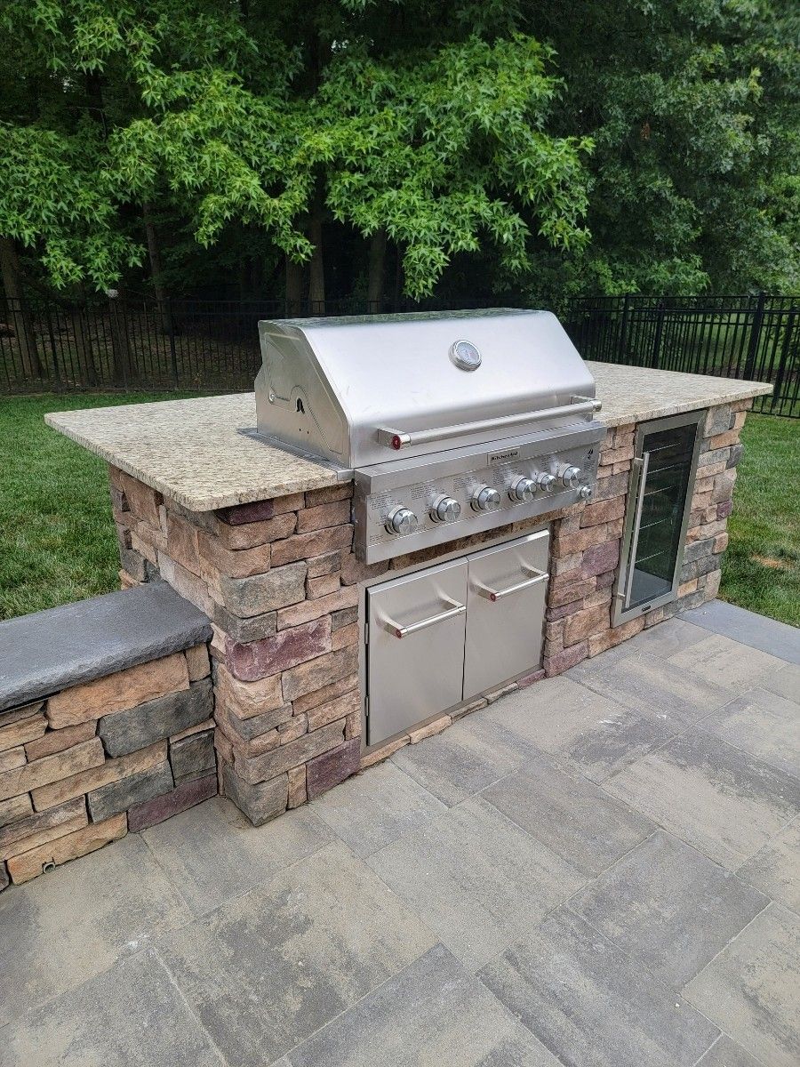 Outdoor kitchen with stone facing, stainless steel grill, countertop, and storage; backyard setting.