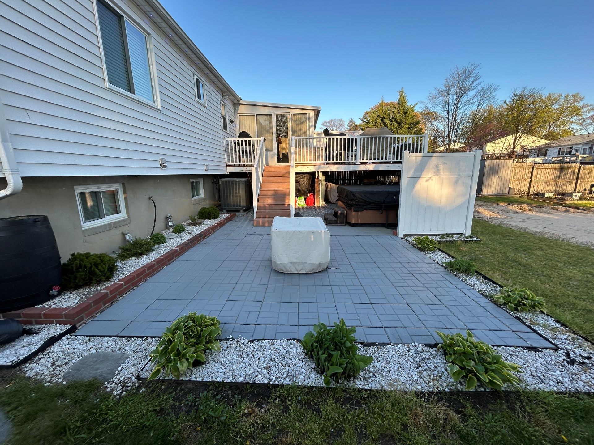 Backyard patio with deck, house, gray pavers, white rock border, plants, and a covered fire pit.