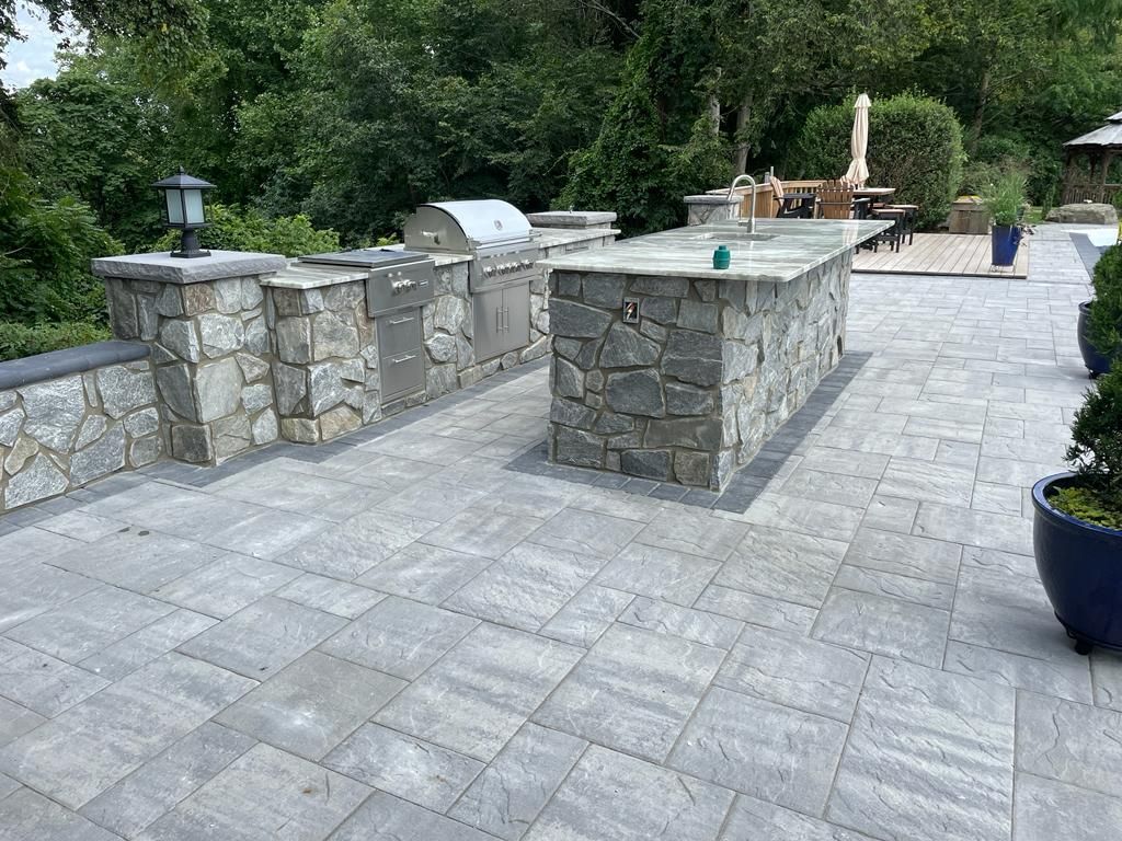 Outdoor kitchen with stone counters and grill on a stone patio with greenery in the background.