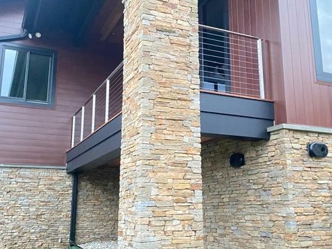 Stone-clad exterior of a house with a balcony featuring cable railing; brown siding and dark trim.