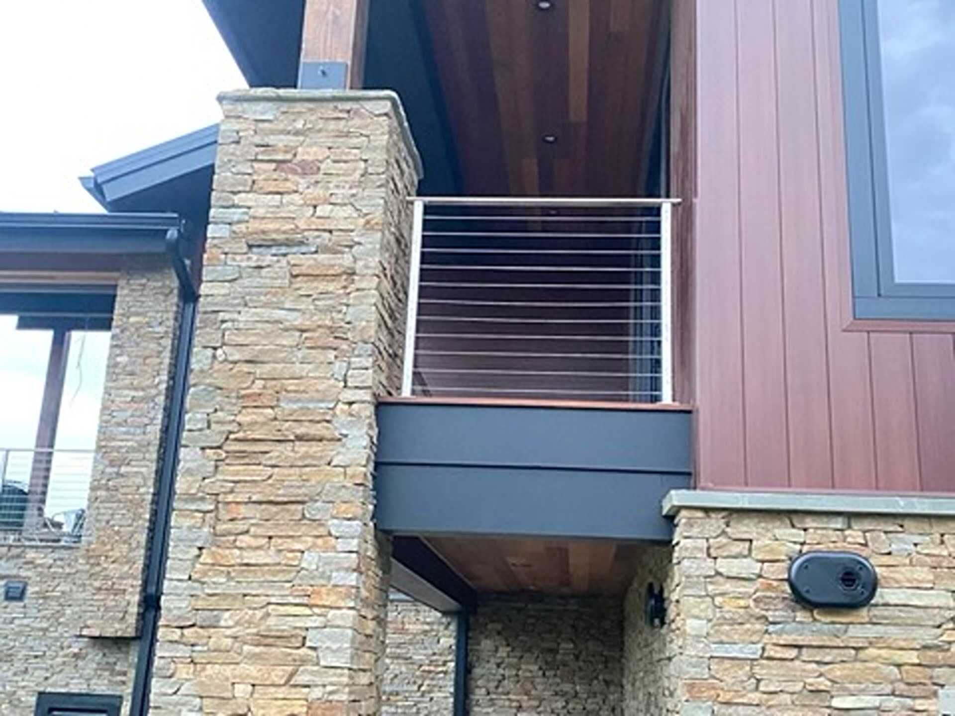 Exterior view of a modern house with stone columns, a deck with horizontal metal railings, and wood paneling.