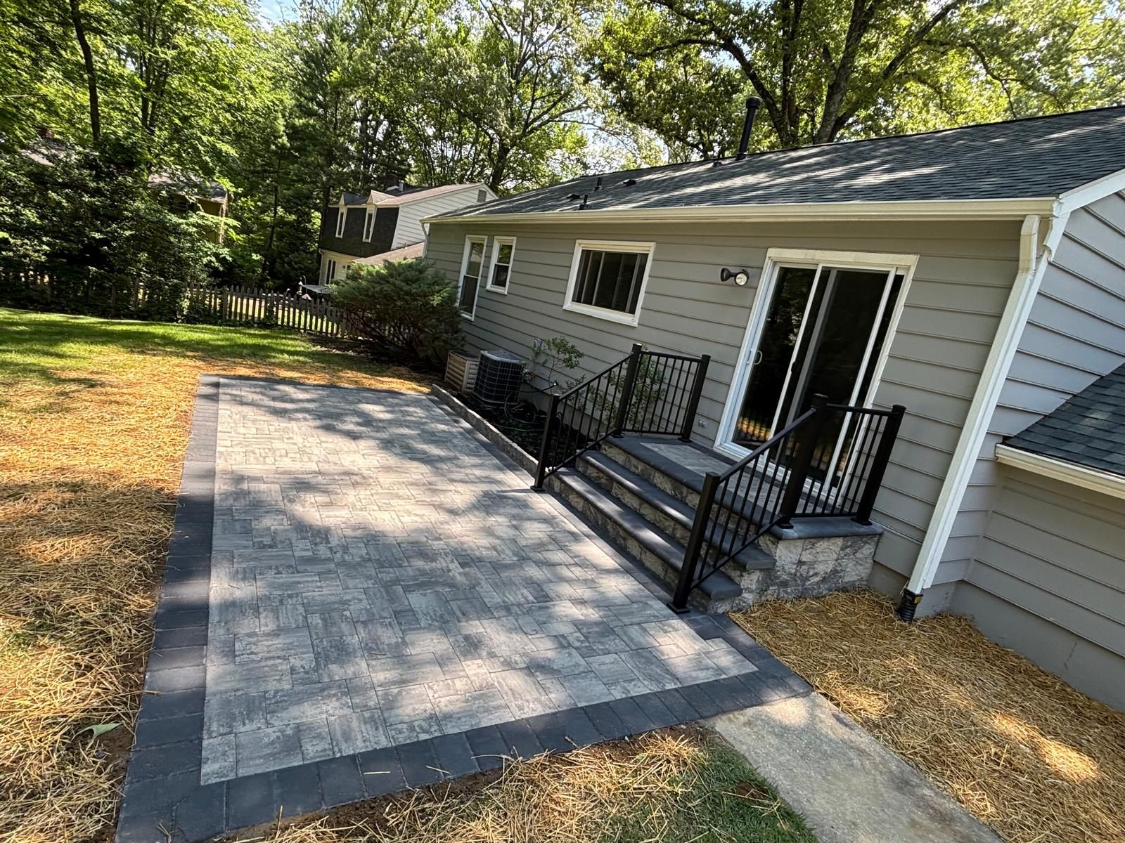 Gray patio with dark border, stairs, sliding glass door, and black railings on a house with a yard.