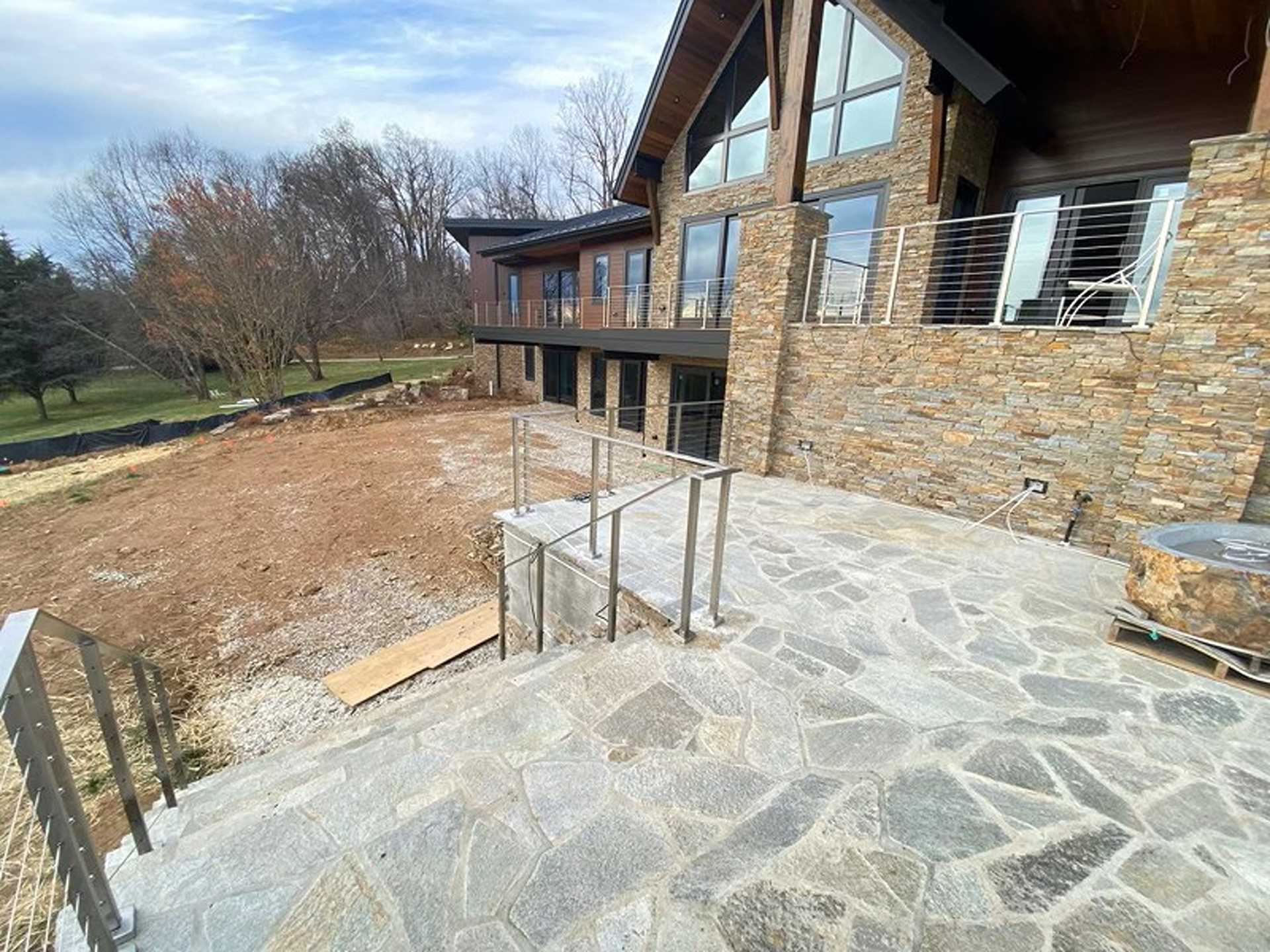 Stone patio with steps leading to a modern house with stone facade and upper-level balcony. Brown, grey, and blue hues.