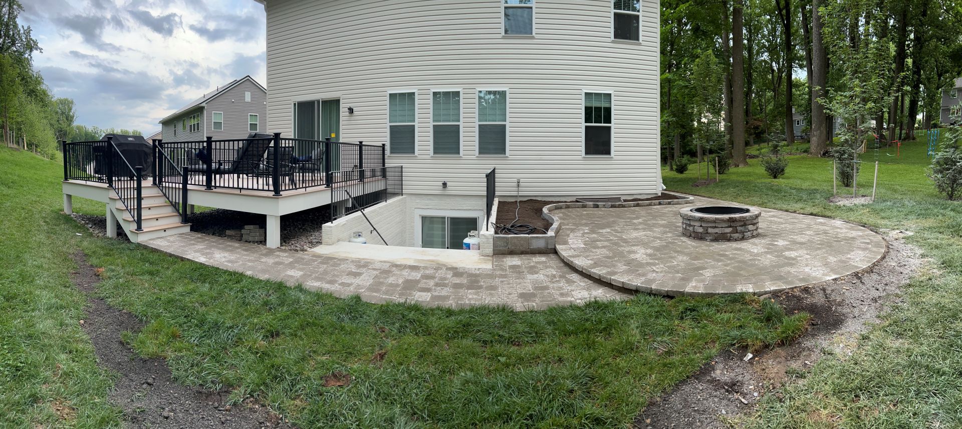 Backyard of a house with a deck, patio, and fire pit on a grassy area, surrounded by trees.