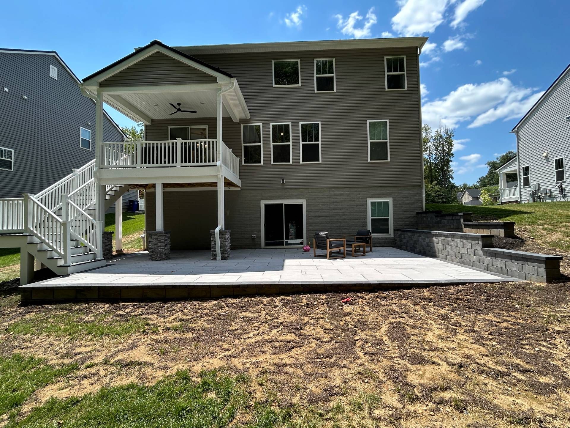 Back of a two-story house with a covered deck, patio, and tiered retaining walls. Blue sky background.