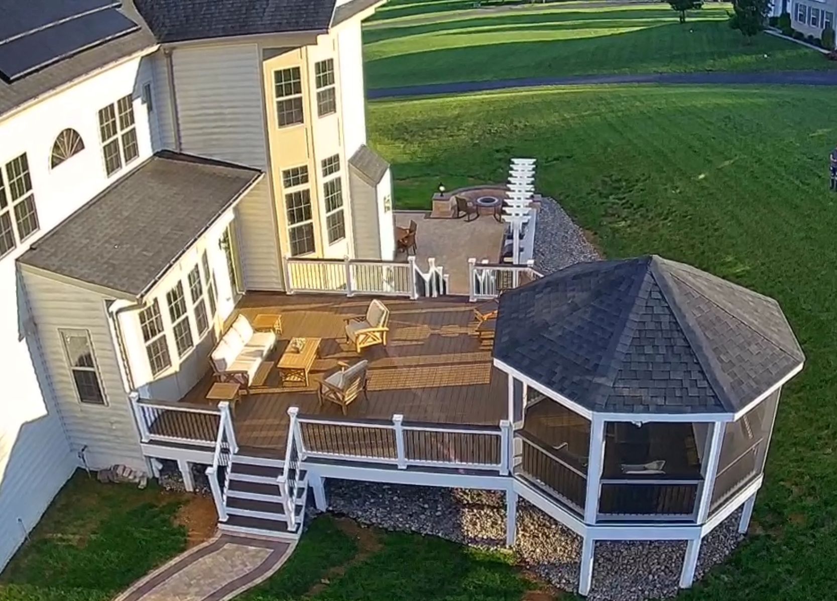 Aerial view of a home with a deck and gazebo. Deck has seating. Gazebo has screen walls. Green lawn surrounds the structures.