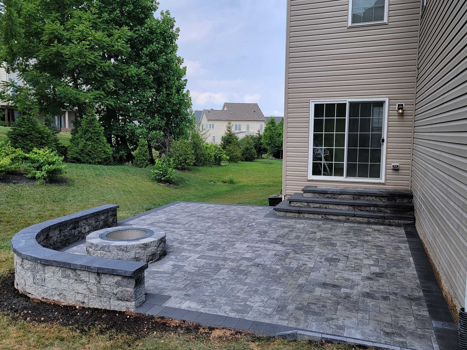 Paver patio with fire pit, steps, and sliding glass door next to a house with beige siding and green grass.